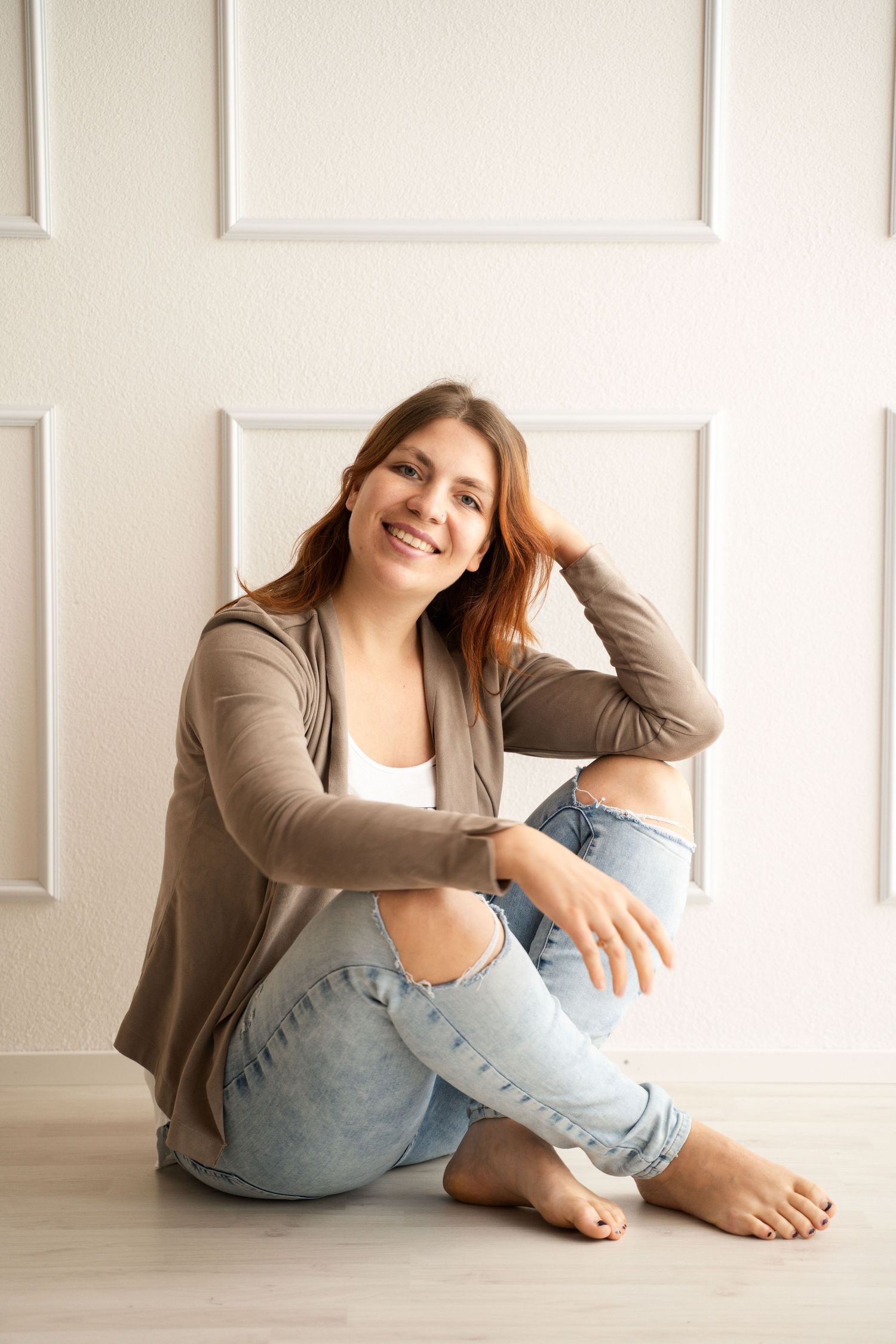 Smiling young woman sitting barefoot on floor in ripped jeans and brown cardigan against white wall.