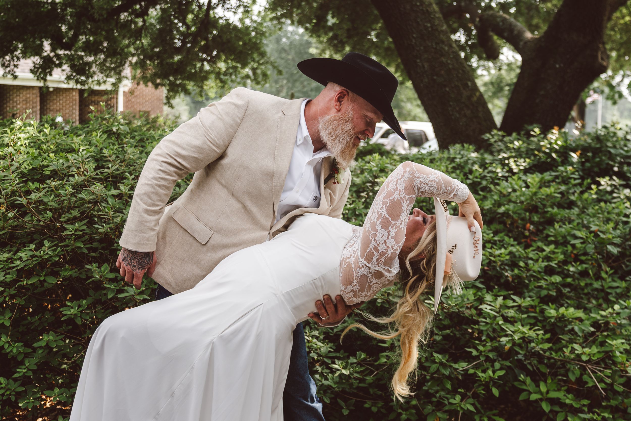 Groom in tan suit and black cowboy hat dips bride in white lace dress outdoors among green bushes.