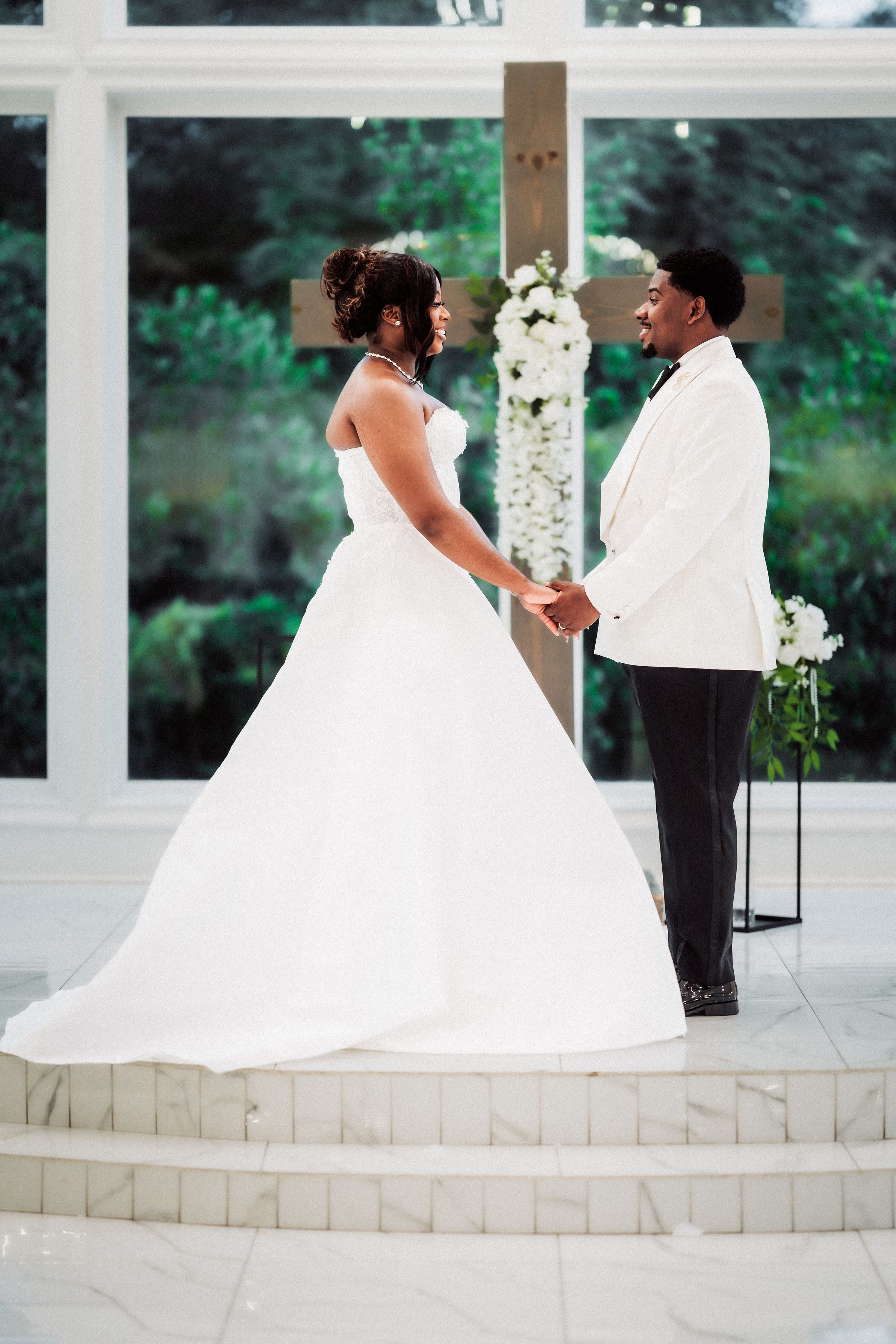 A bride and groom hold hands at the altar in a sunlit chapel, she in a white ball gown, he in a white tuxedo jacket.