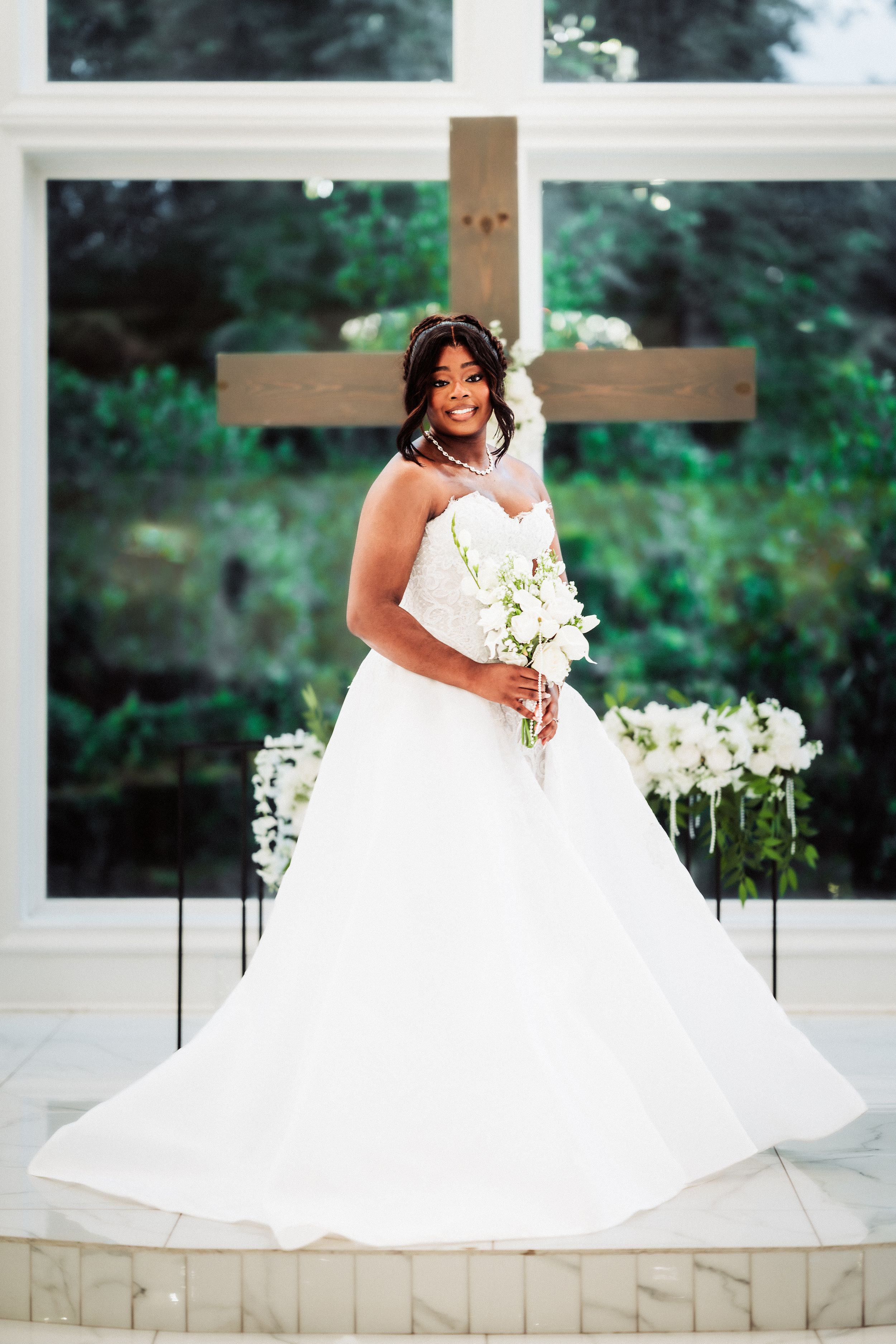 Bride in white ball gown holding bouquet stands before wooden cross in sunlit chapel.