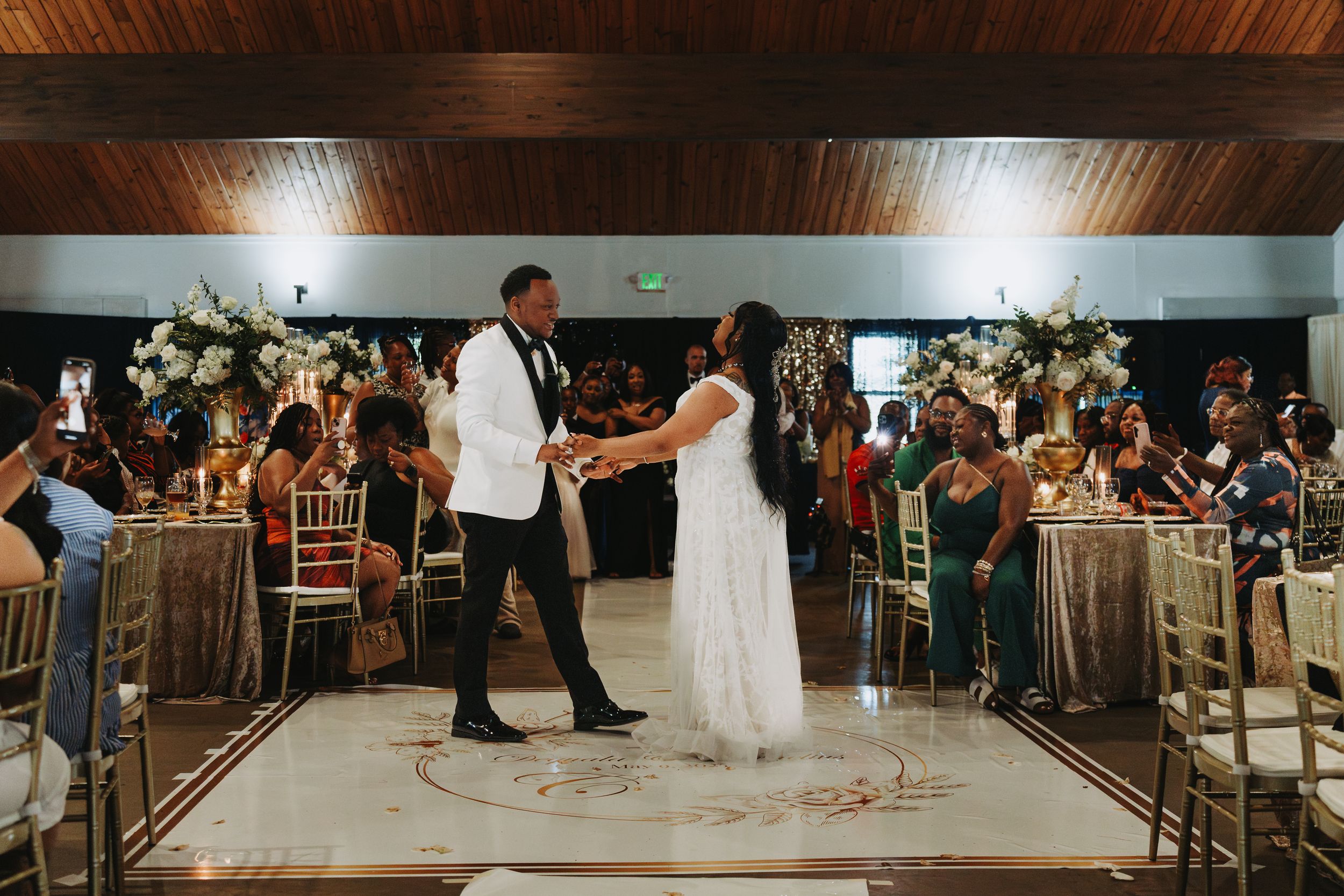 Wedding guests watch in anticipation as a couple practices their first dance in a rustic wood-paneled venue.