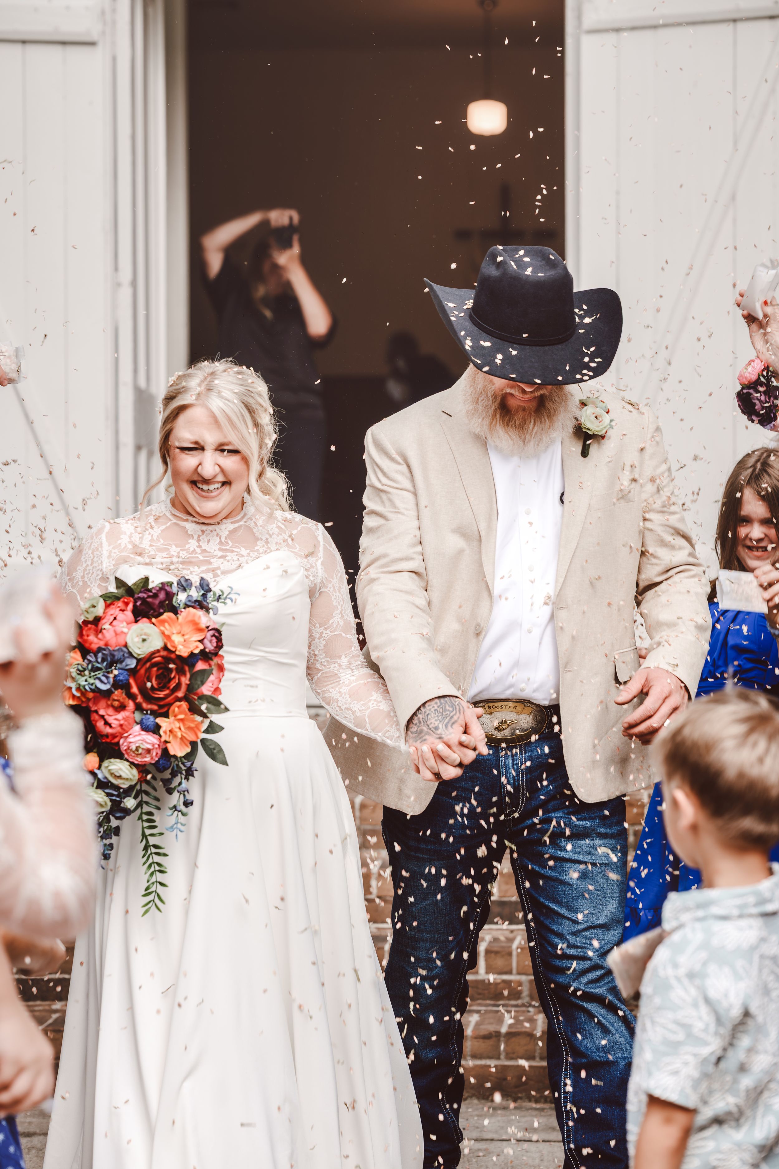Bride and groom exit church in confetti shower, she holds colorful bouquet, he wears cowboy hat and tan blazer.