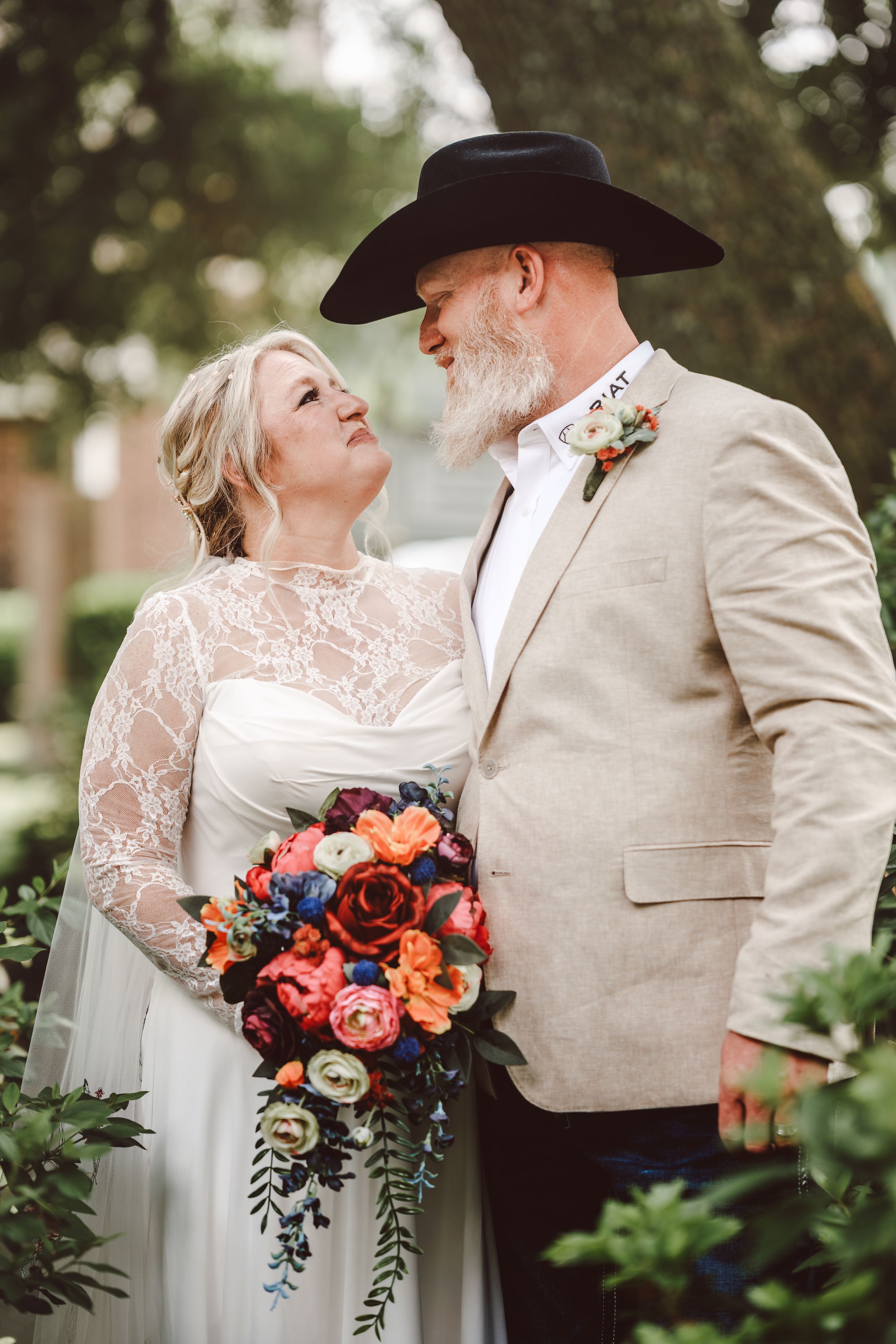 Couple in wedding attire, bride holding colorful bouquet, groom in cowboy hat and tan blazer, gazing at each other outdoors.