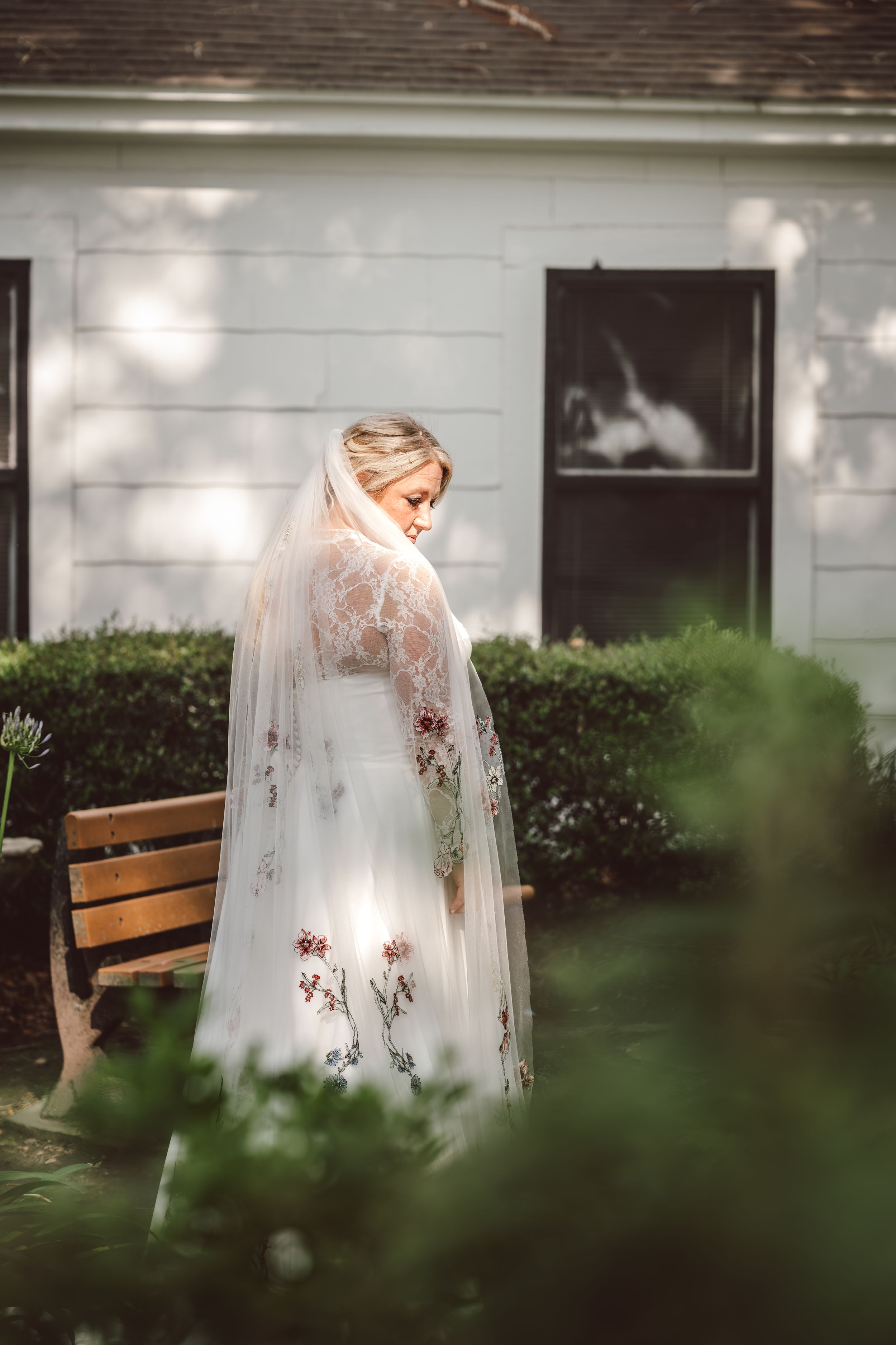 Bride in floral embroidered wedding dress and lace veil standing in garden near wooden bench.