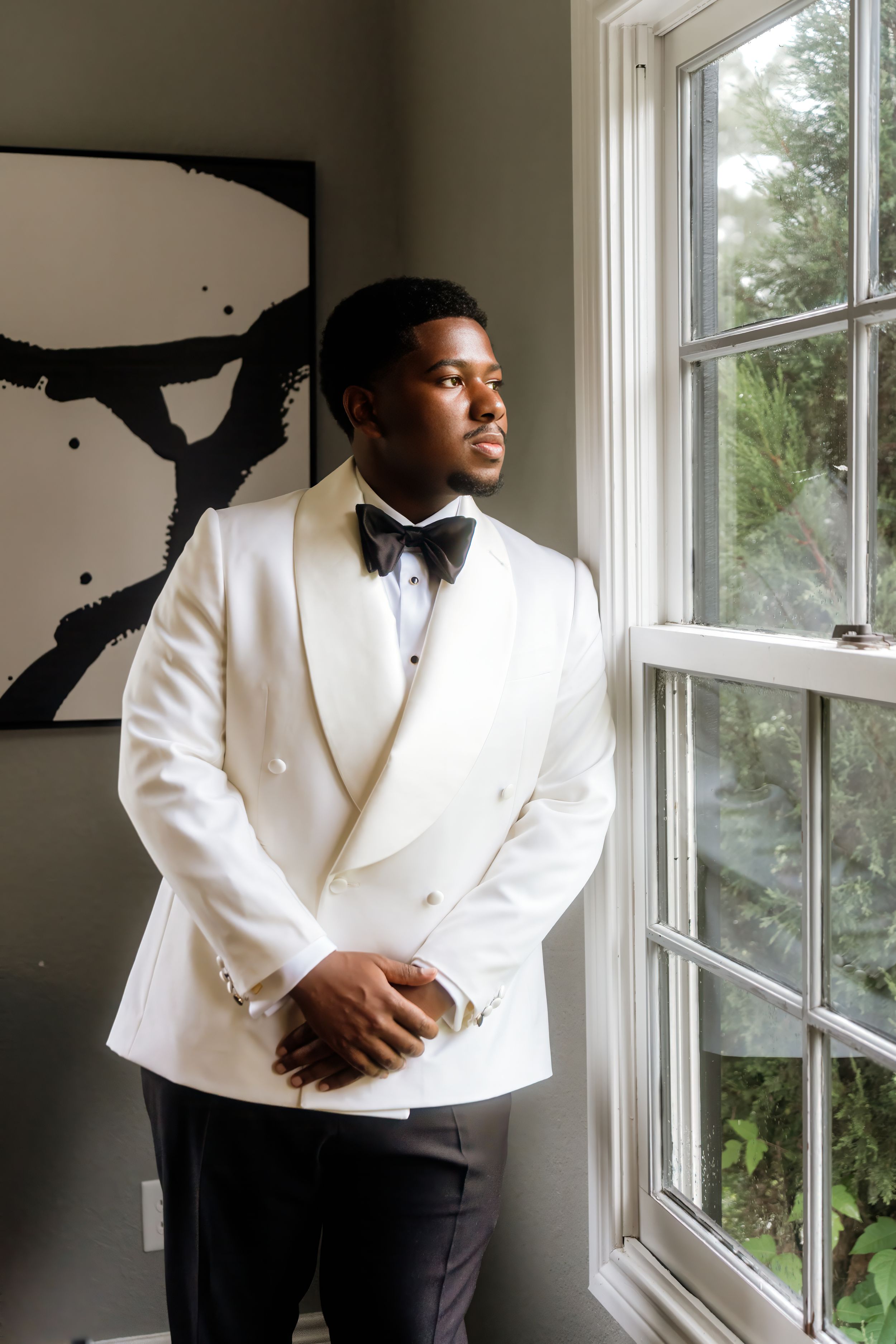 A groom in a white double-breasted tuxedo jacket and black bow tie stands by a window gazing outside.