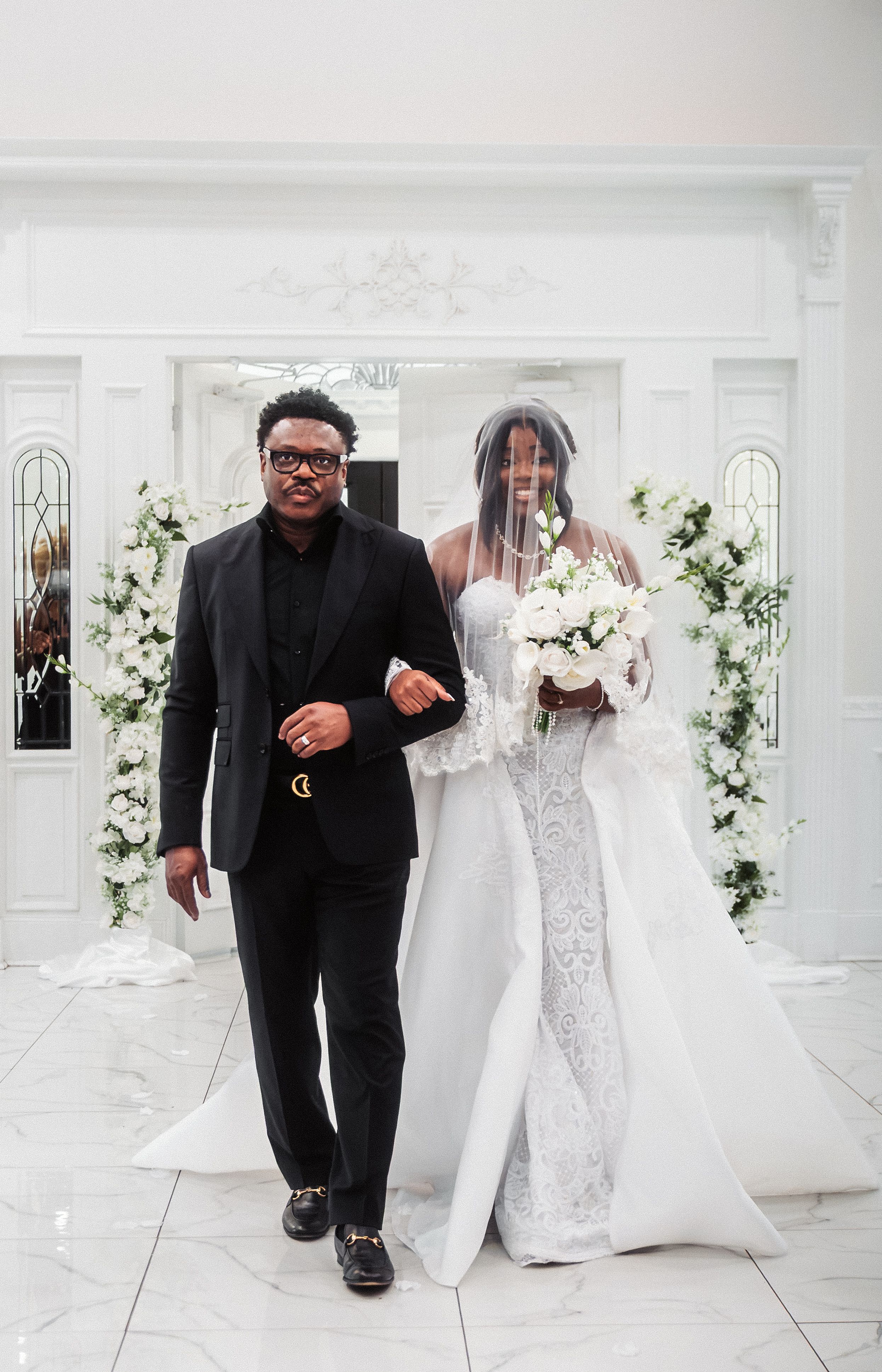 Bride in white lace gown holding bouquet walks with man in black suit at elegant wedding ceremony.