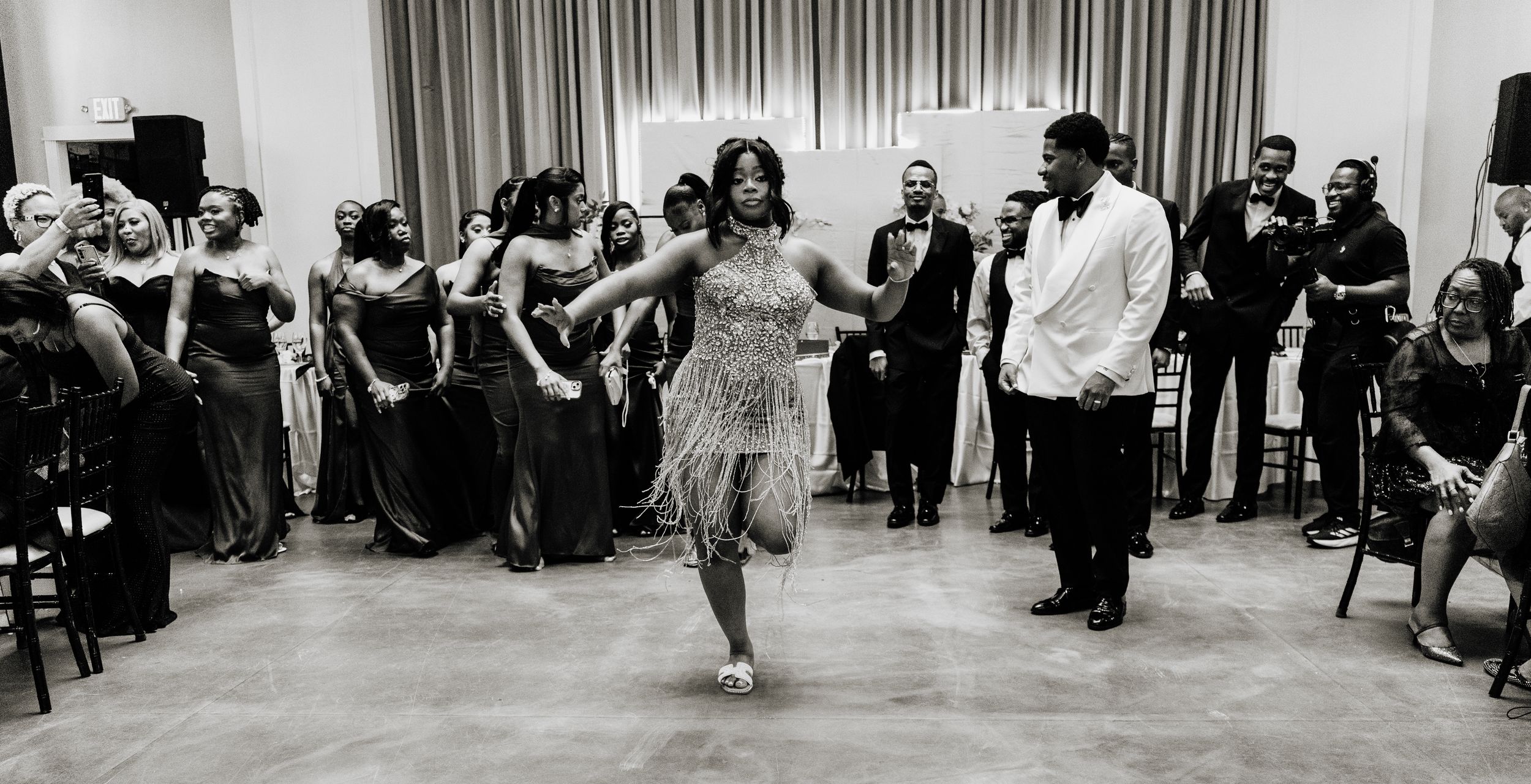 Black and white photo of a woman in a fringed dress dancing at a formal event with guests watching.