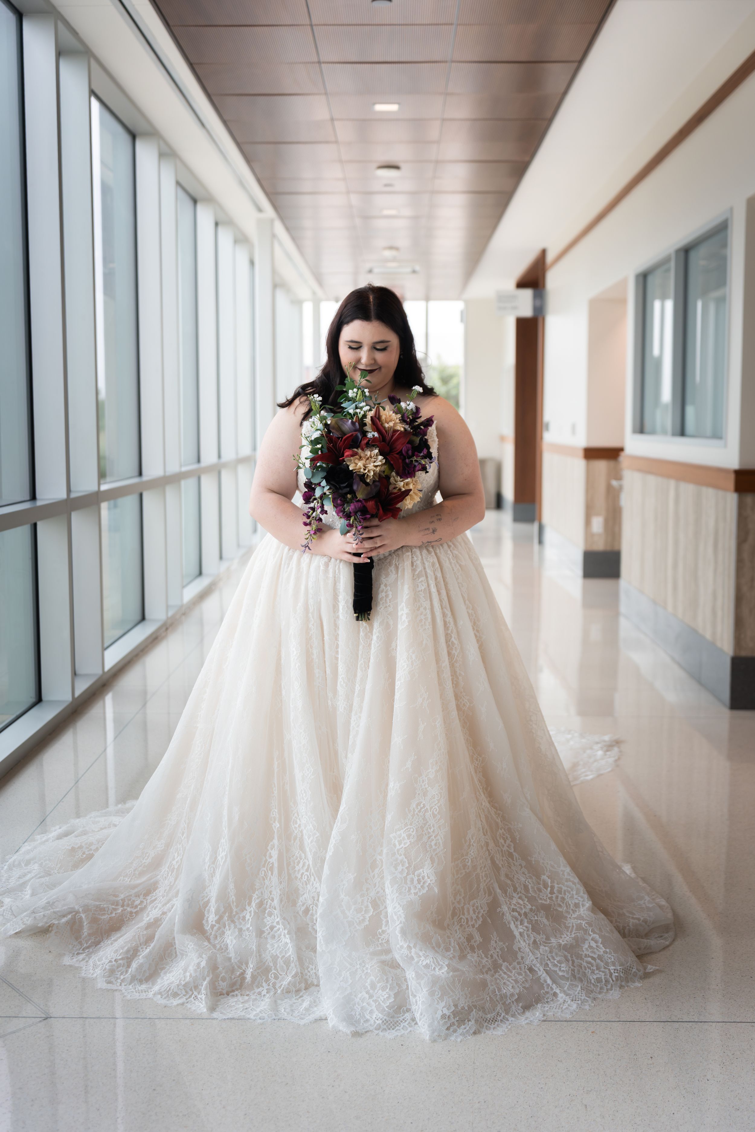 Bride in lace ballgown holding burgundy bouquet stands in sunlit corridor.