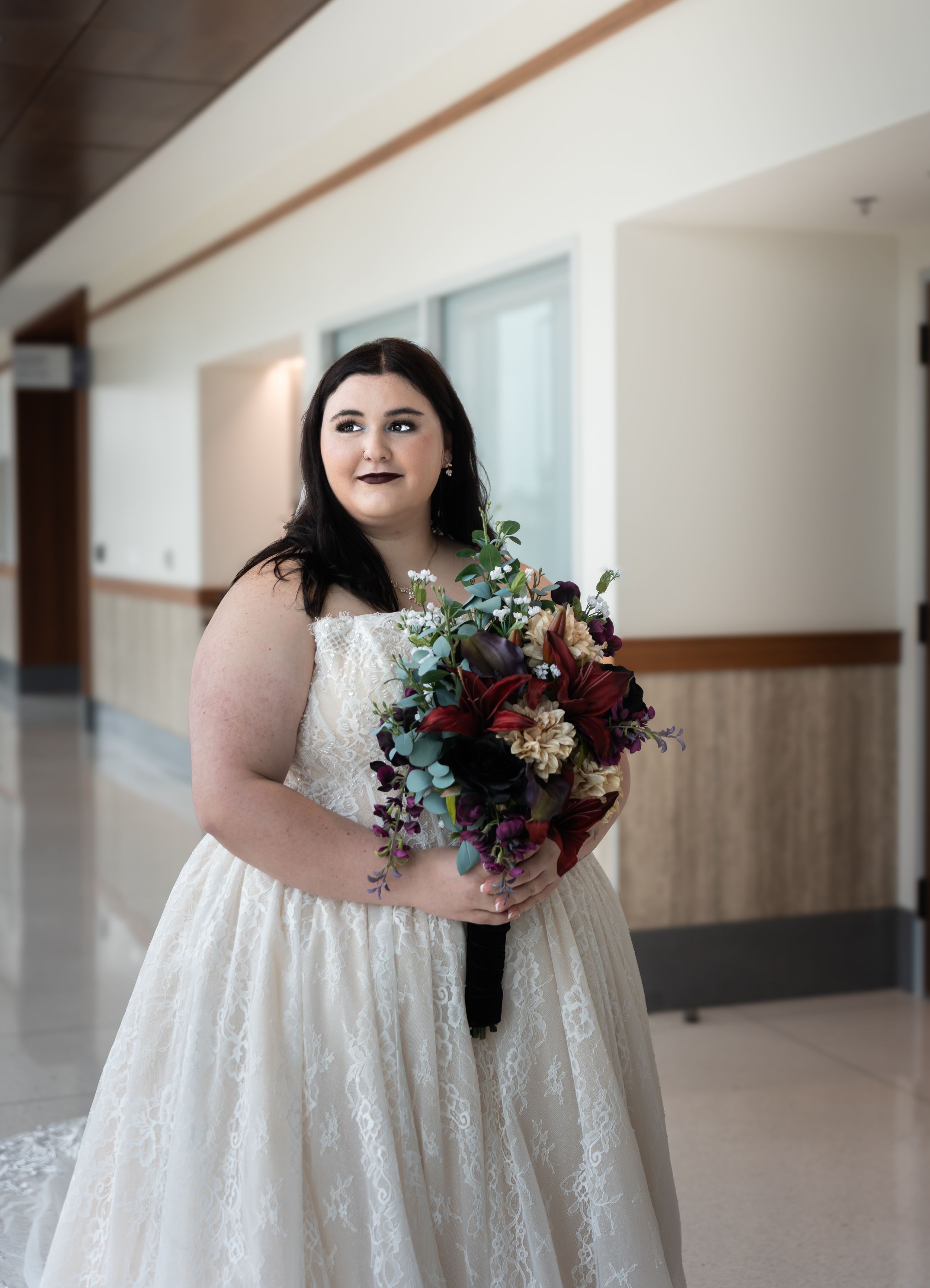 Bride in white lace gown holds dark floral bouquet with burgundy blooms and greenery in elegant indoor setting.