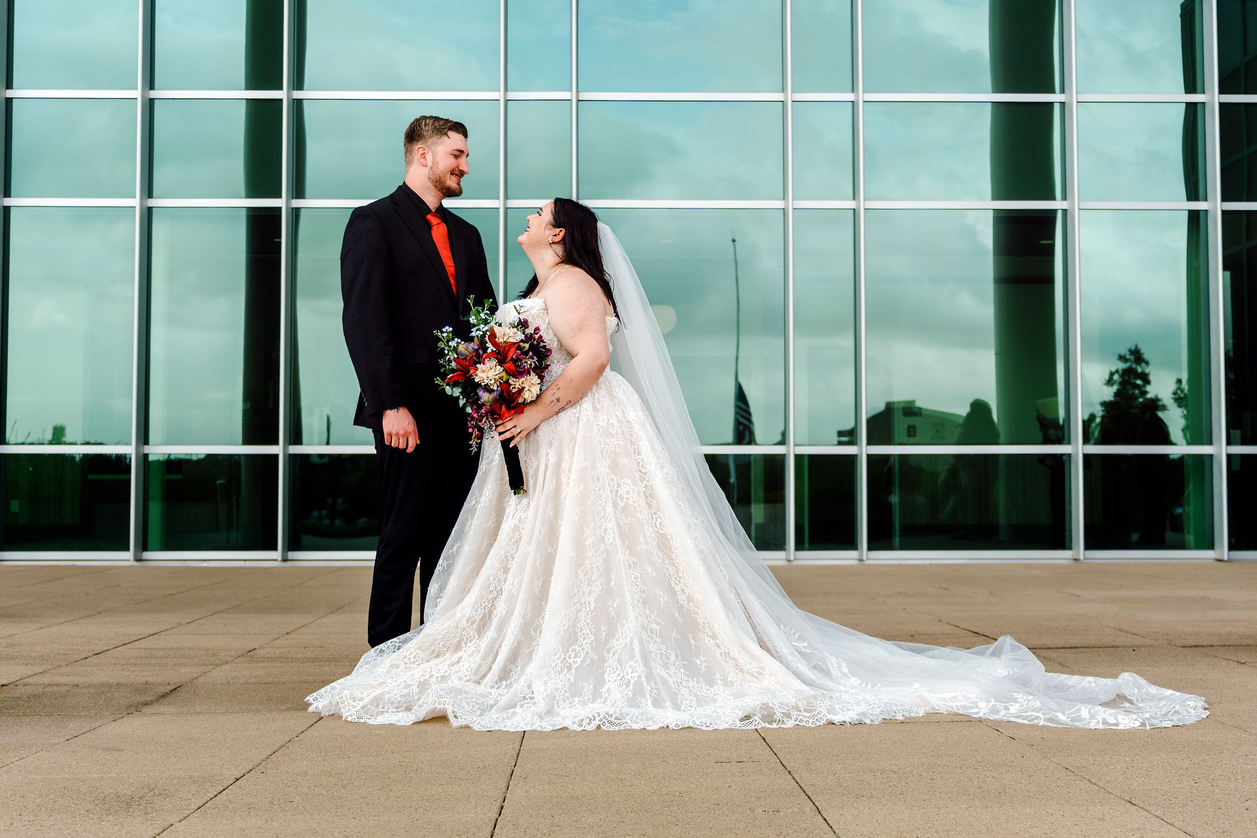 Bride in lace gown with bouquet and groom in black suit with red tie pose outside glass building.