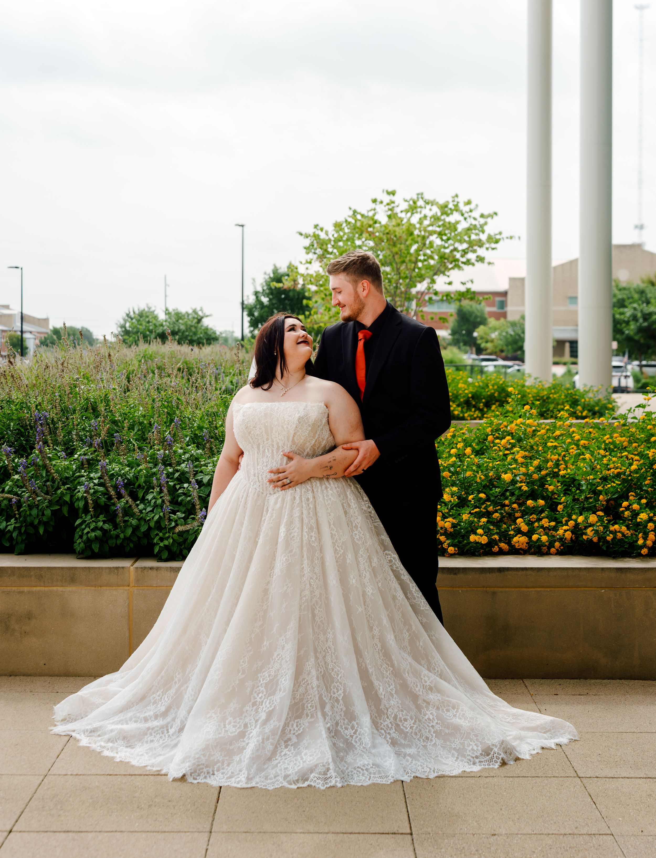 Bride in lace ball gown and groom in black suit with red tie share a romantic moment outdoors near floral gardens.