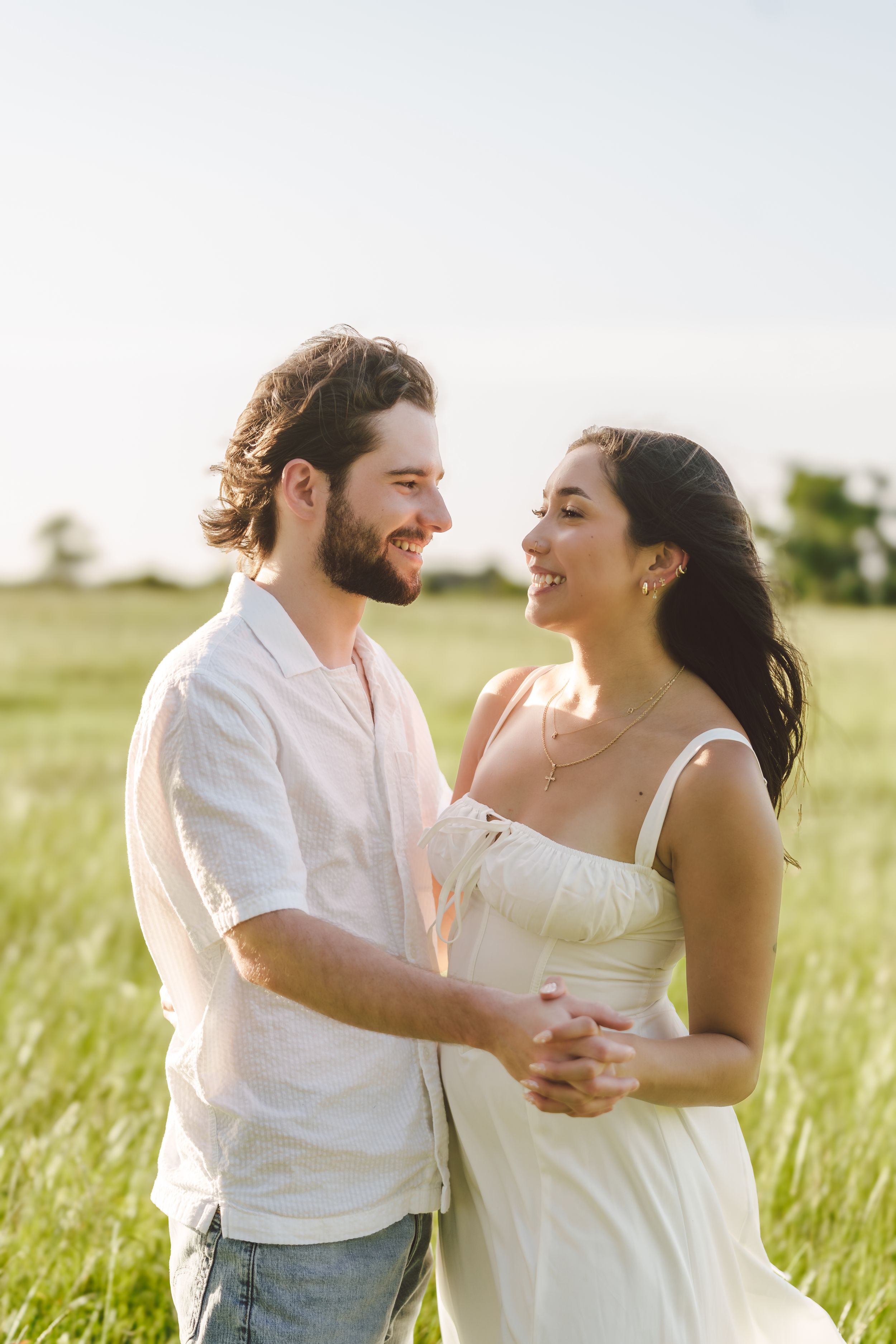 Couple in white dresses dance and embrace joyfully in a sunlit meadow with tall grass.