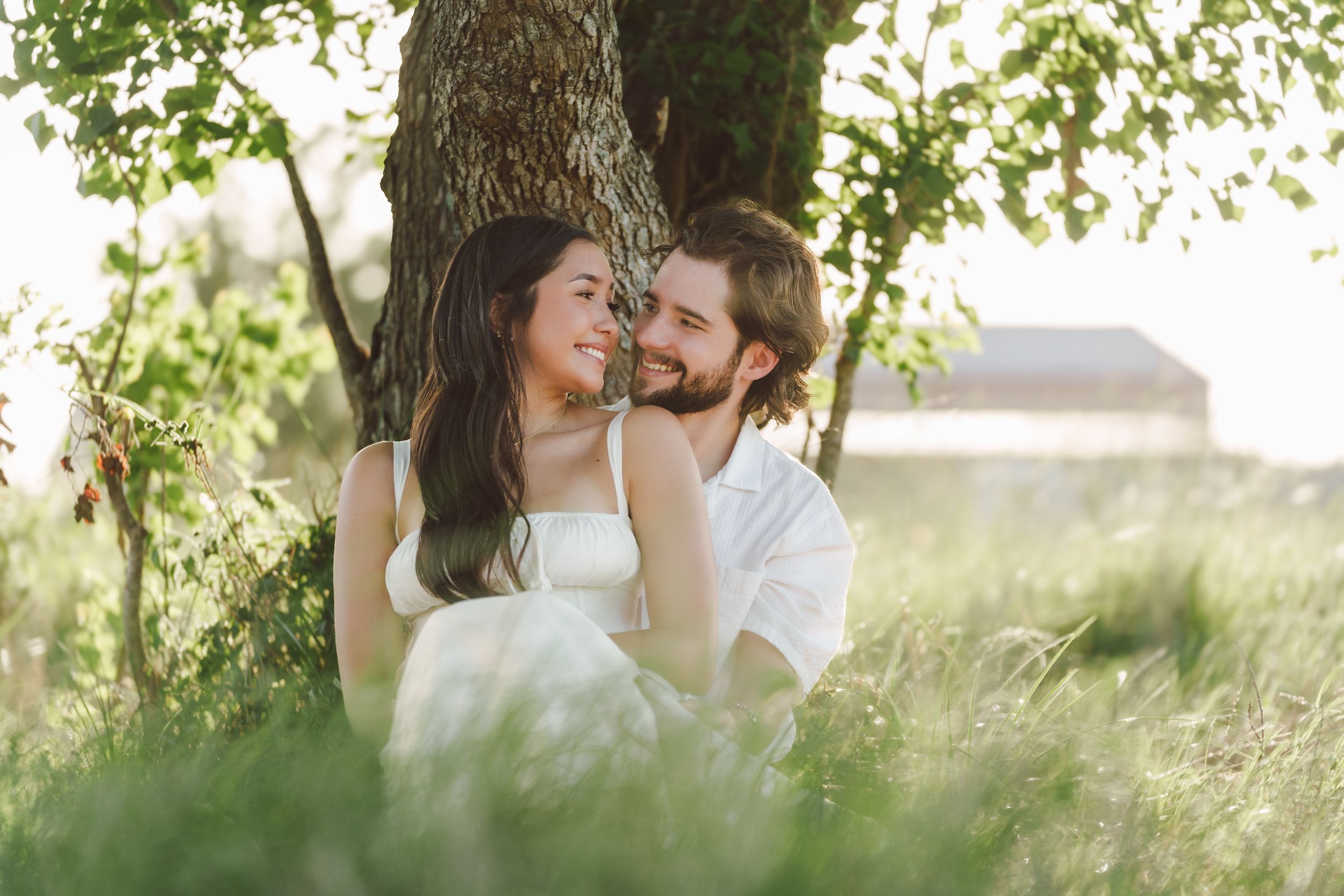 Two people sharing a tender moment together while sitting in tall grass near a tree during golden hour.