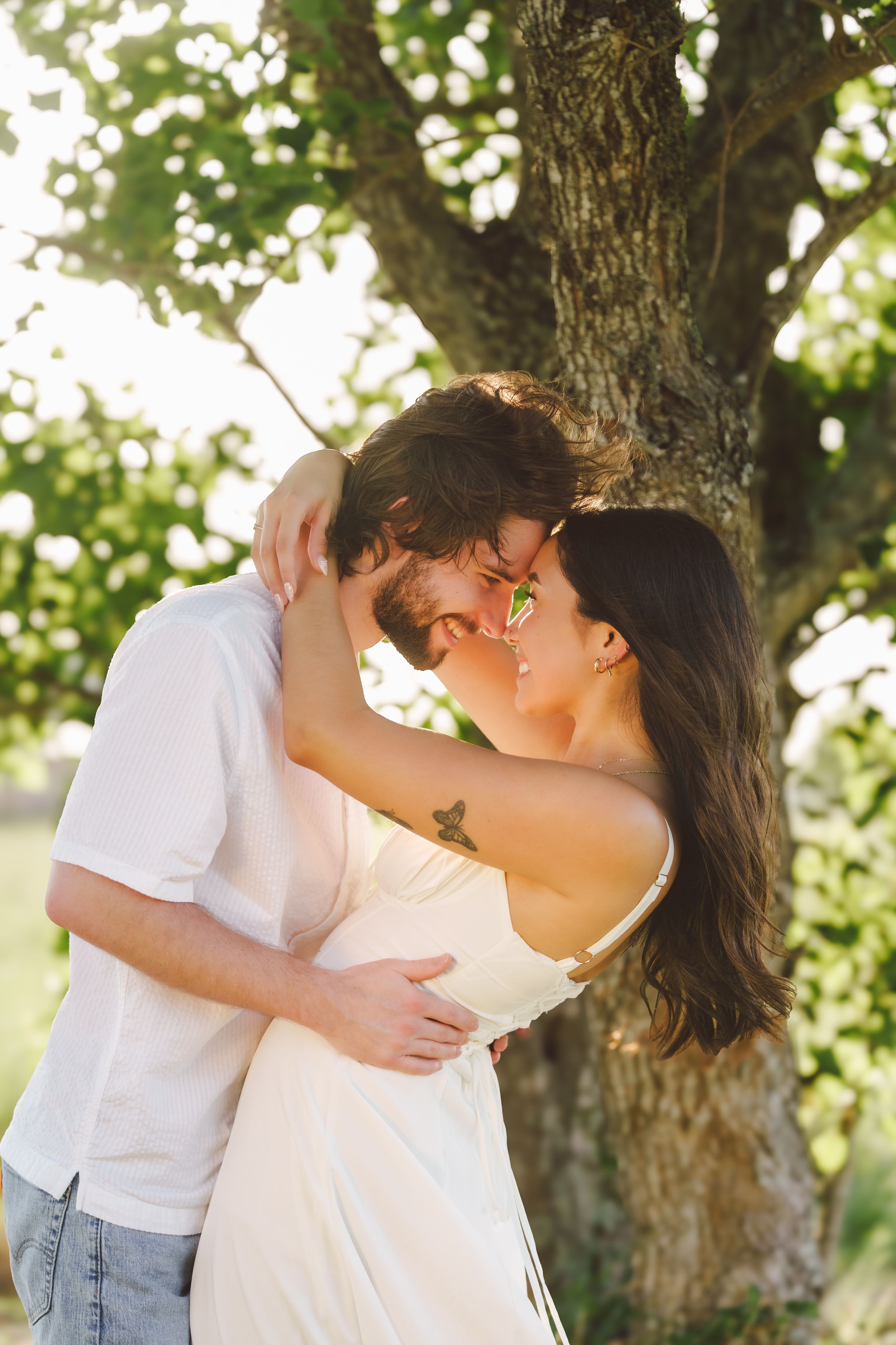 Couple shares intimate moments beneath a large tree during a summer photoshoot.