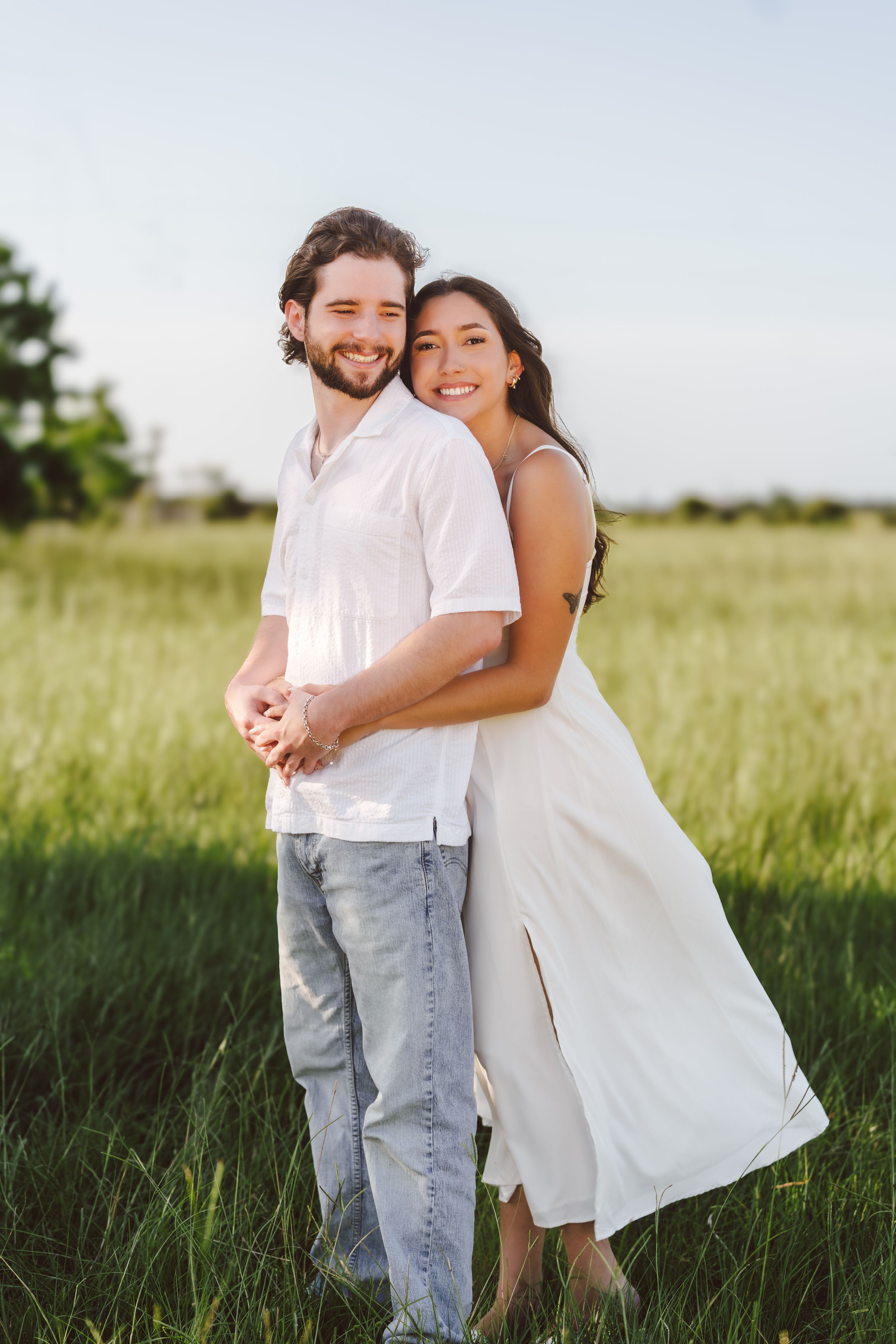 A couple stands together embracing in a sunlit grassy field, with her white dress flowing in the breeze.