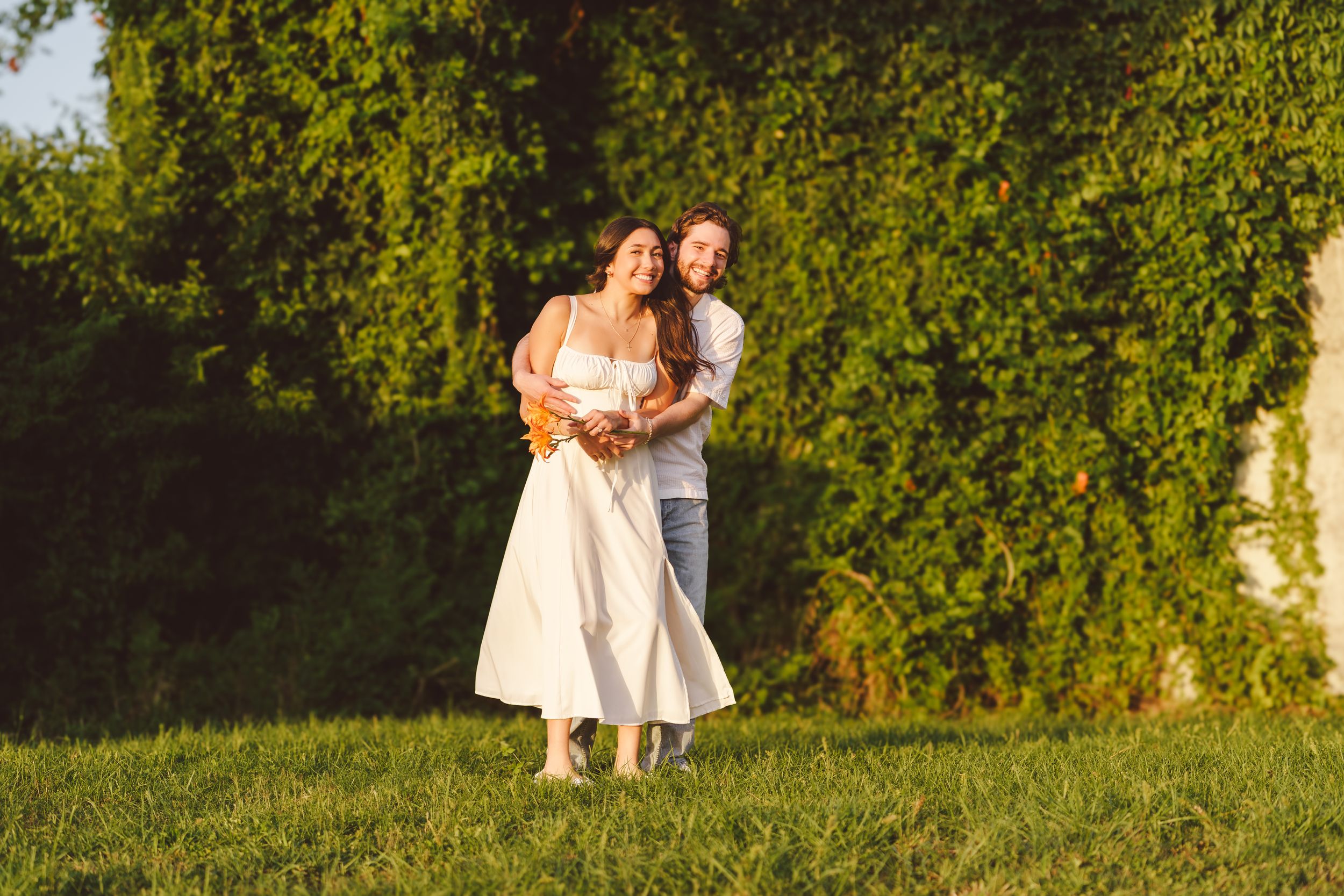 A couple embraces while standing together on a grassy hillside with lush green trees in background.