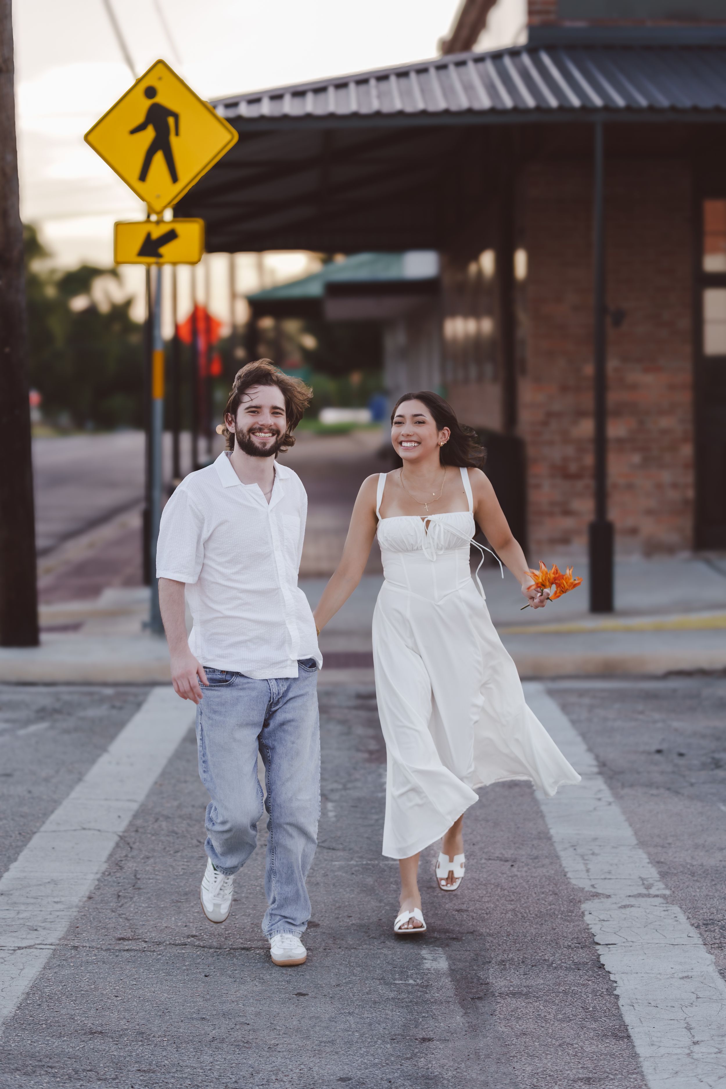 A couple runs playfully across a crosswalk in white outfits near pedestrian signs.