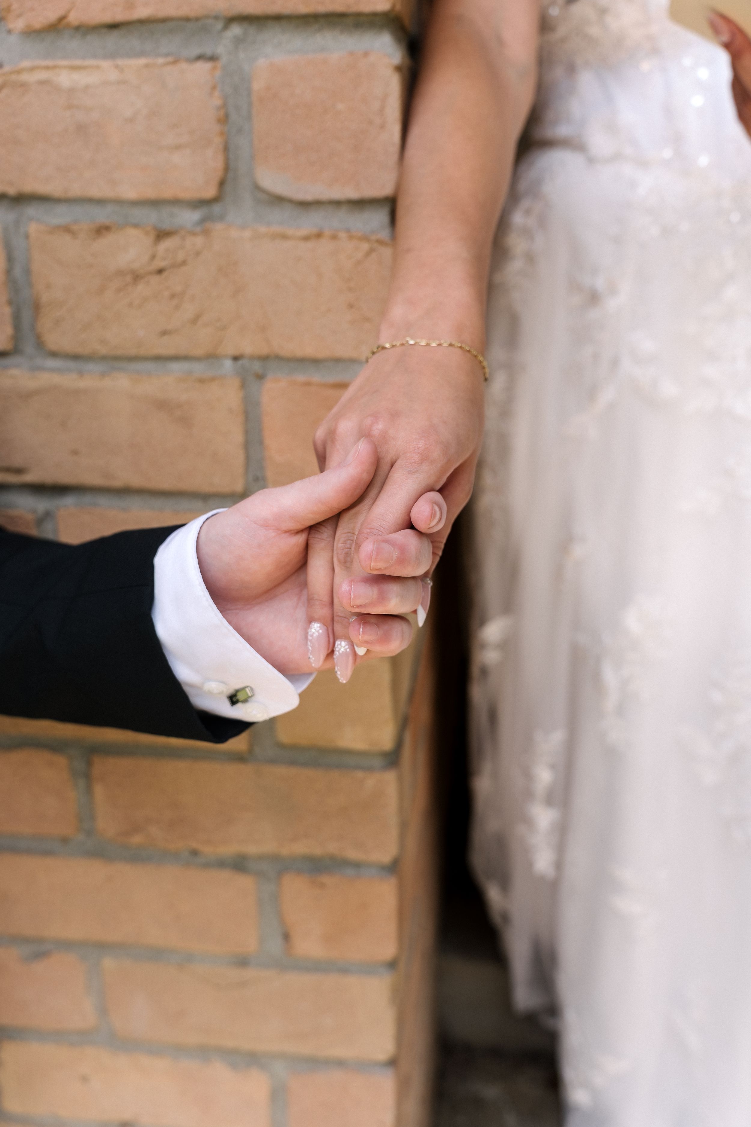 Close-up of a bride and groom holding hands against a brick wall background at their wedding ceremony.