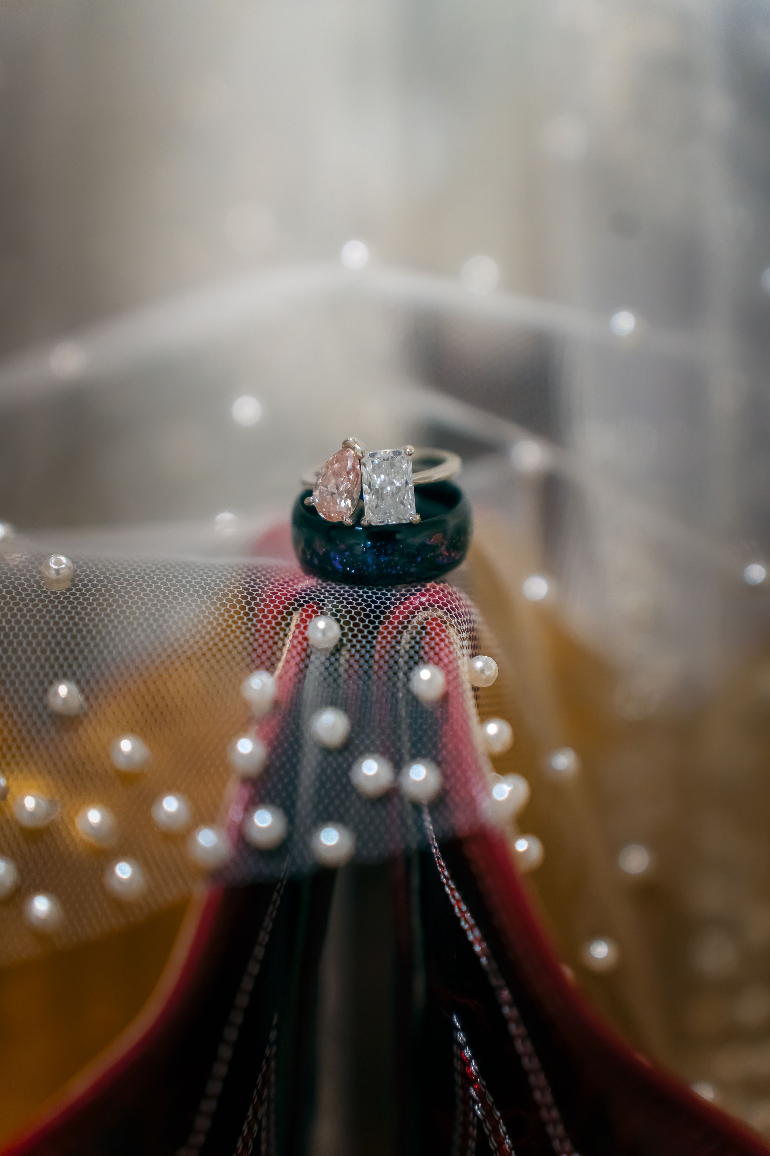 Wedding rings stacked on a dark velvet ring holder, surrounded by a pearl-adorned bridal veil with soft bokeh background.