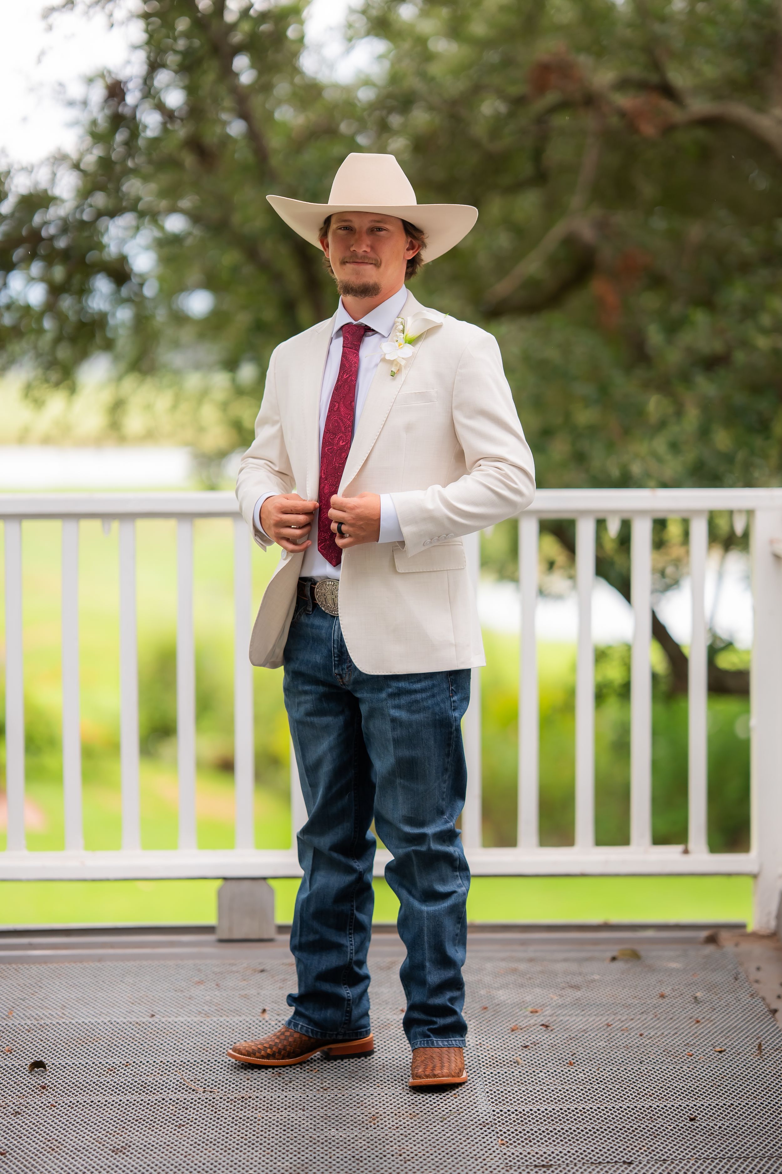 Groom in white blazer, cowboy hat, jeans, and boots with boutonniere stands on deck at outdoor wedding.