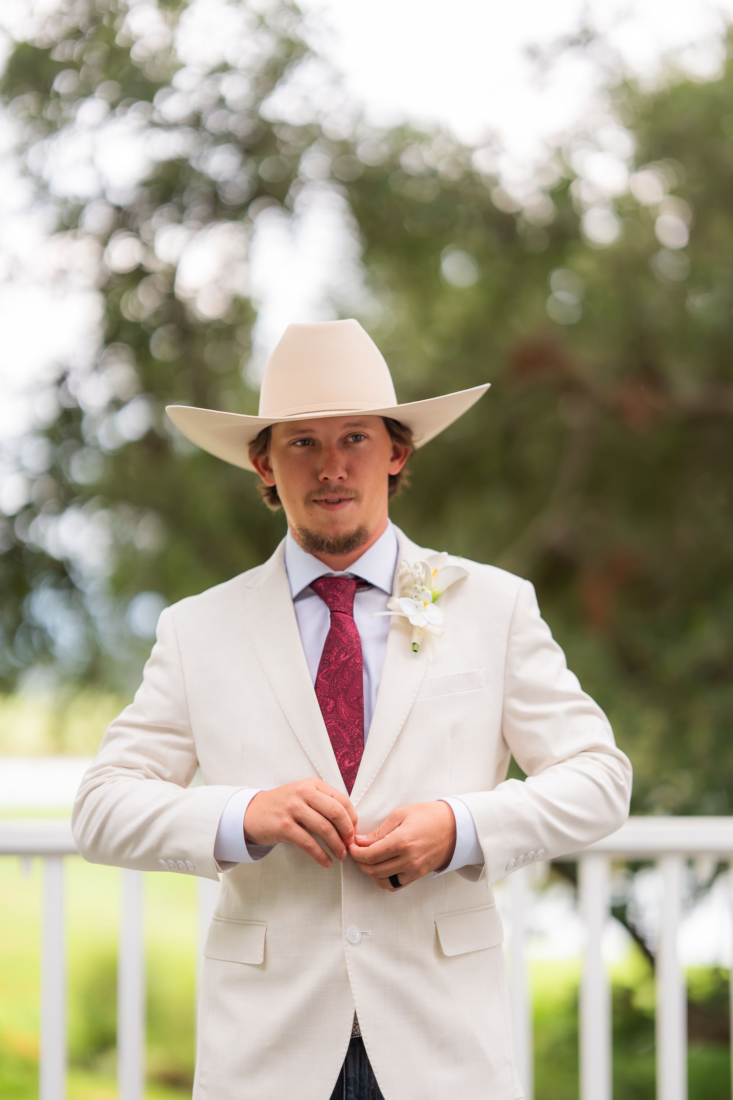 Groom in white suit, red tie, boutonniere, and cowboy hat buttoning jacket outdoors at wedding ceremony.