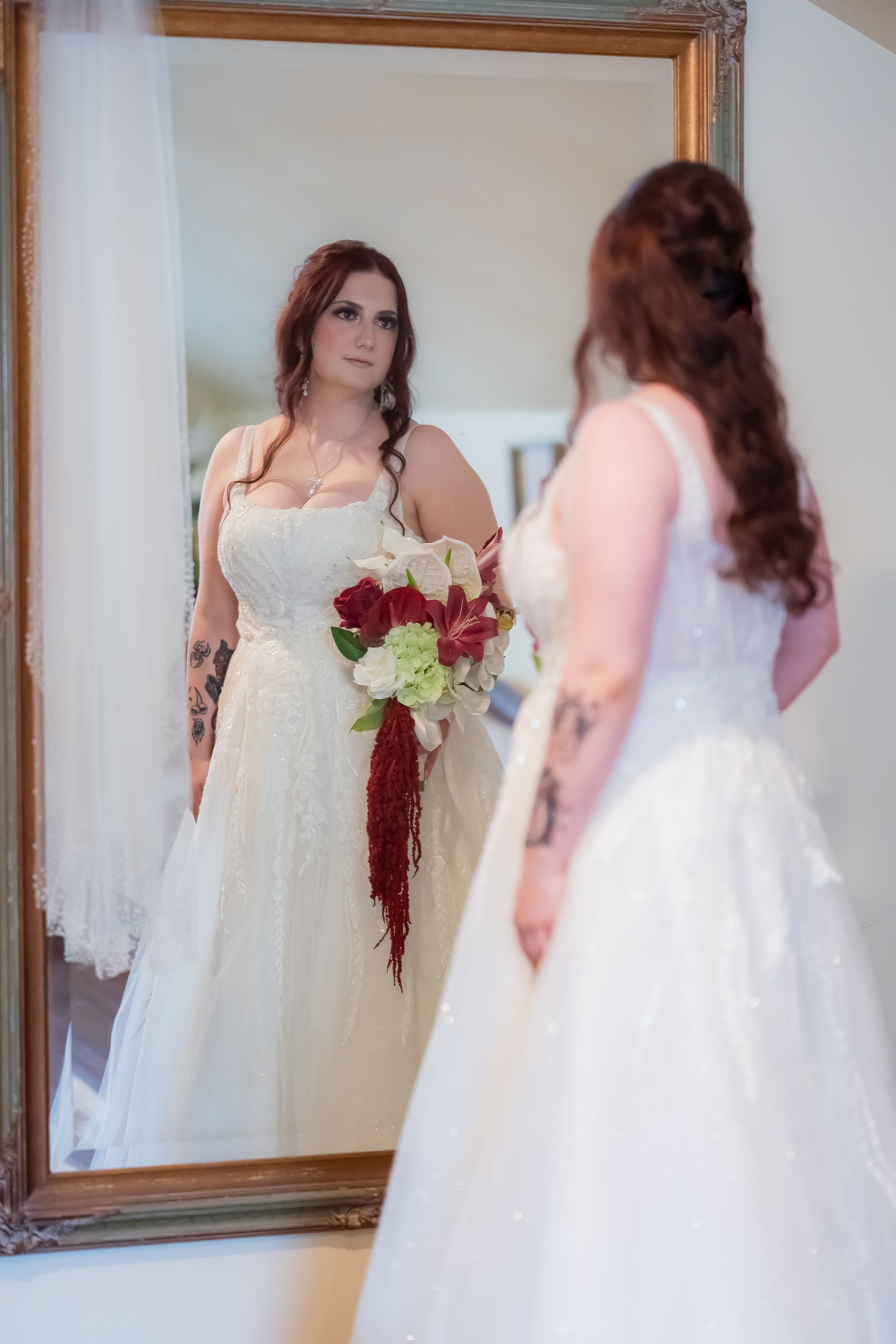 Bride in white lace gown holding red and white bouquet admires her reflection in an ornate gold mirror.