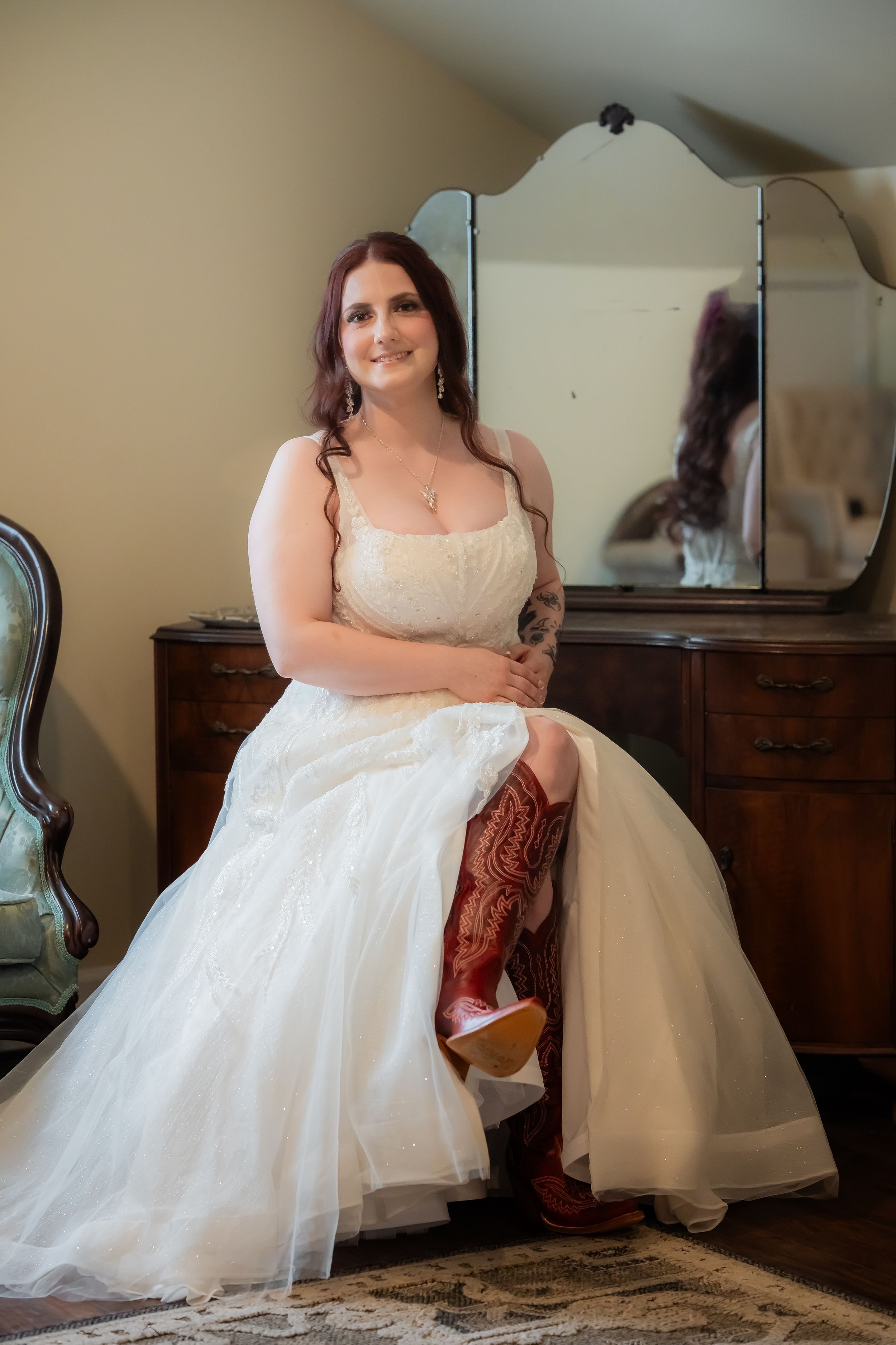 Bride in white wedding gown and red cowboy boots sits before vintage mirror dresser, smiling warmly.