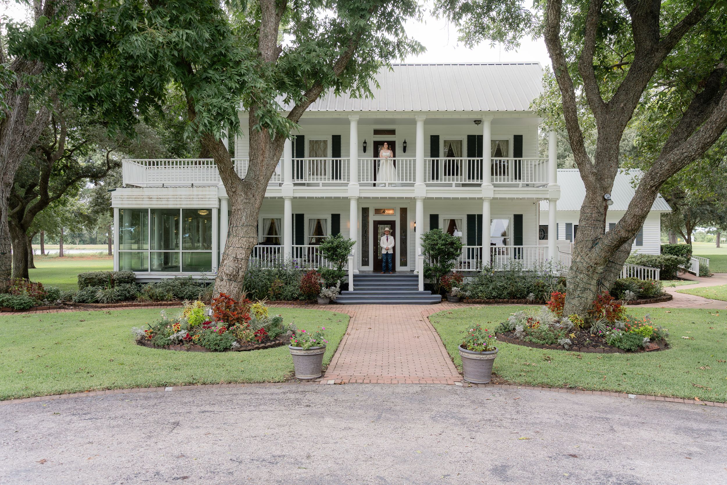 Grand white two-story Southern plantation home with wraparound porches, brick walkway, manicured gardens, and large oak trees.