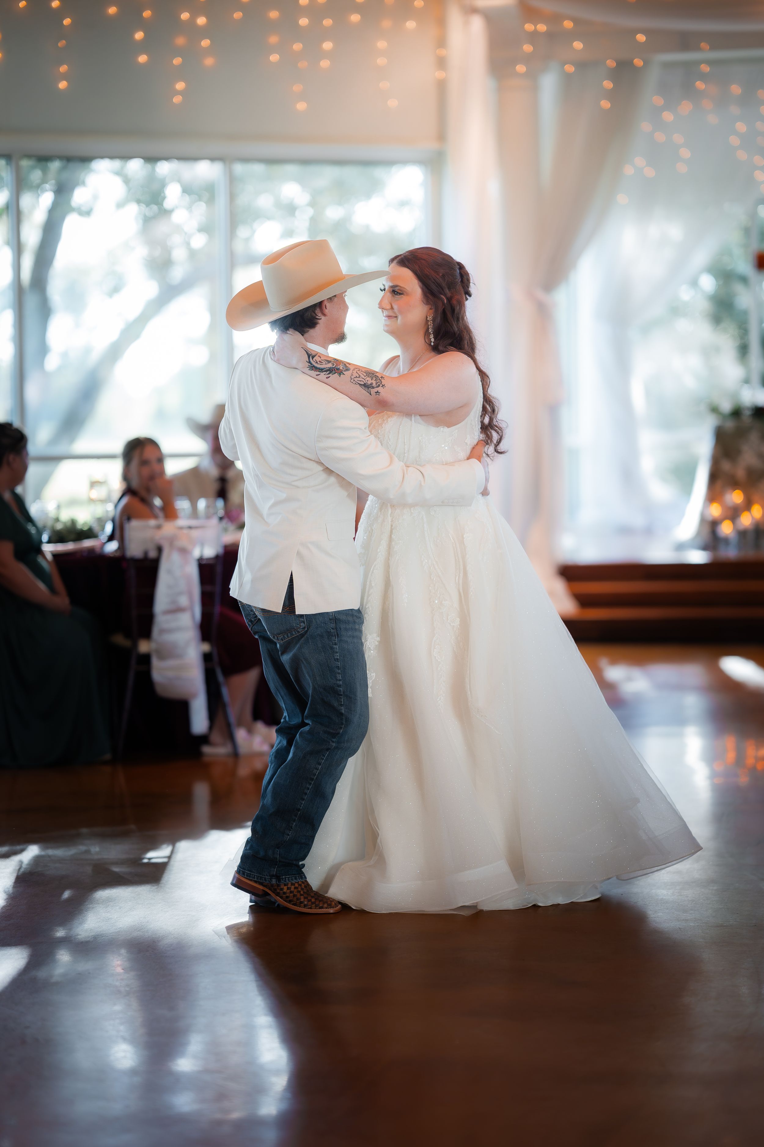 Bride and groom share their first dance at a rustic wedding reception, with string lights and guests in the background.