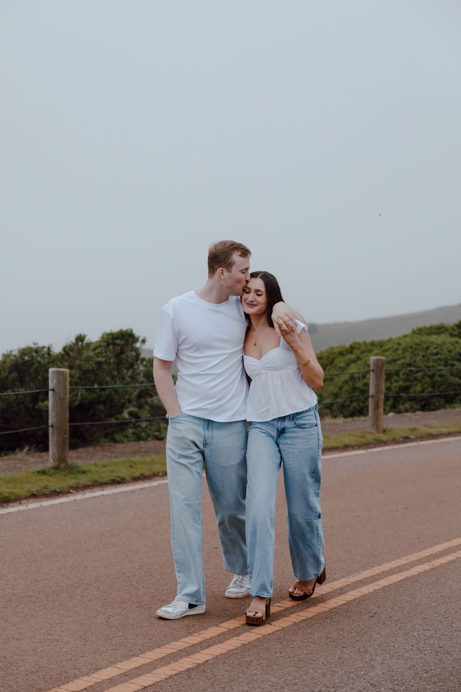 A couple walks together on a scenic coastal path wearing casual denim and white outfits.