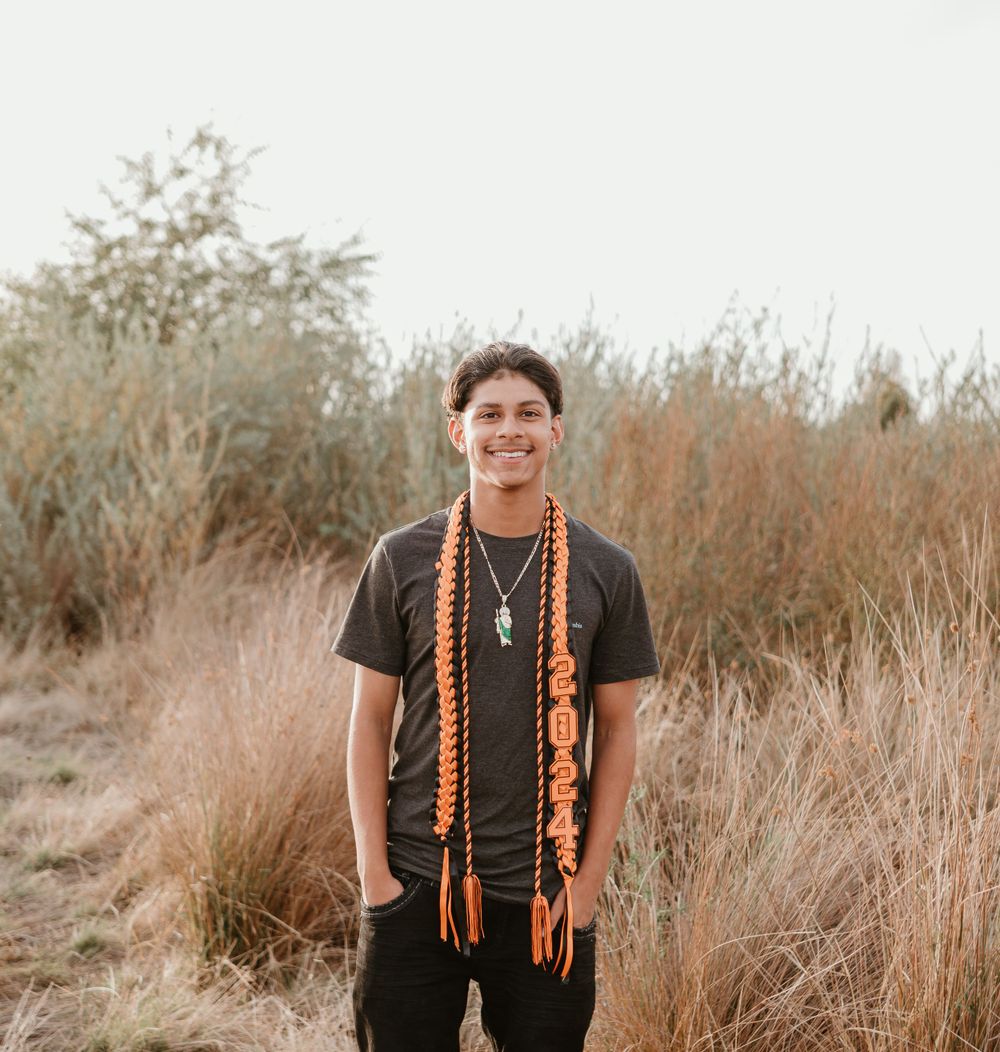 A graduate wearing orange graduation cords stands in a field of tall grass at sunset.