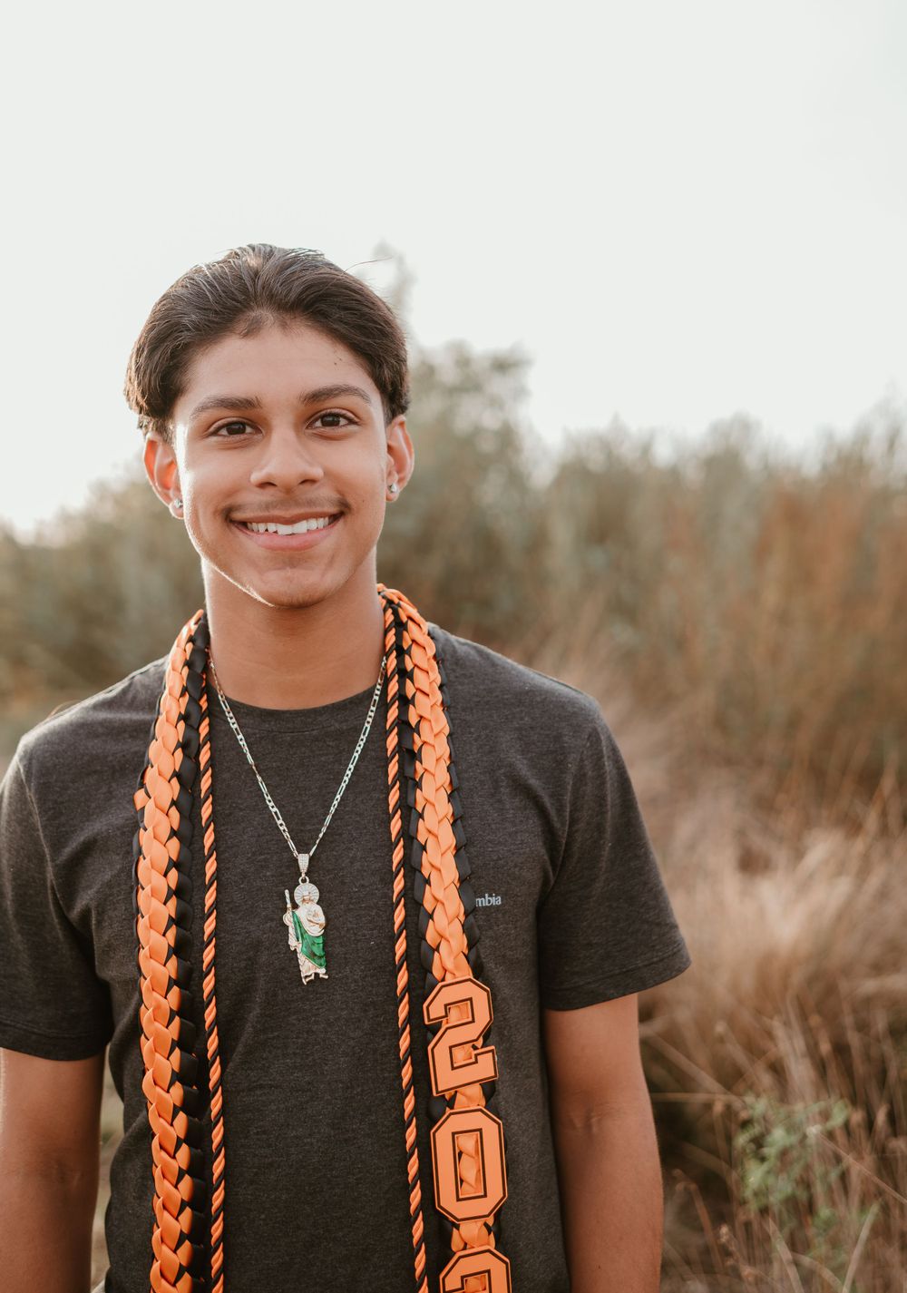 A student wearing orange graduation stoles and a necklace smiles at the camera.