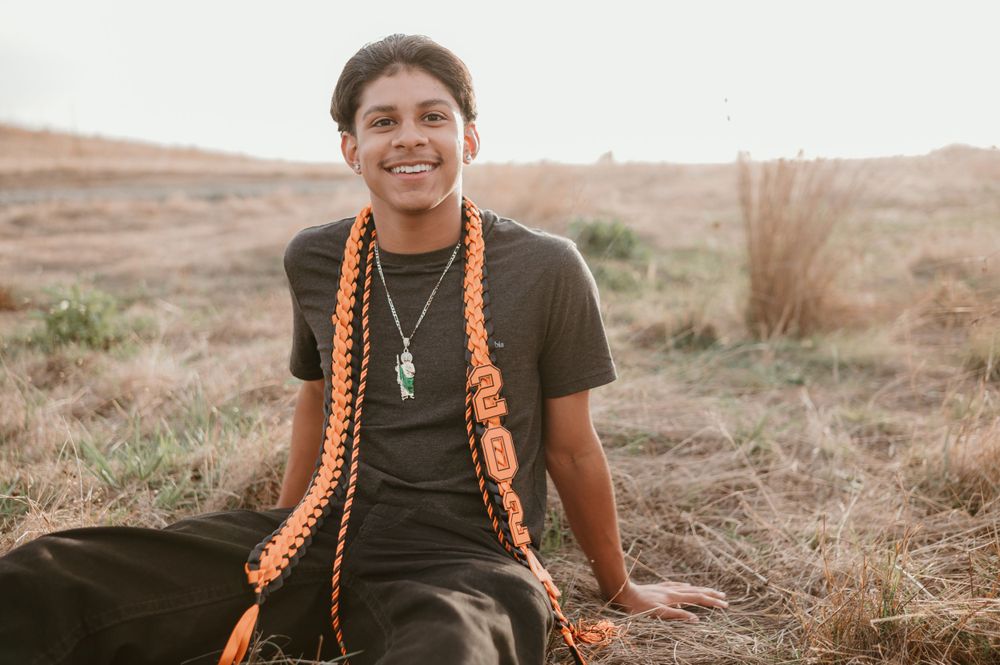 A graduate in a dark t-shirt with orange stoles poses in a natural outdoor setting.