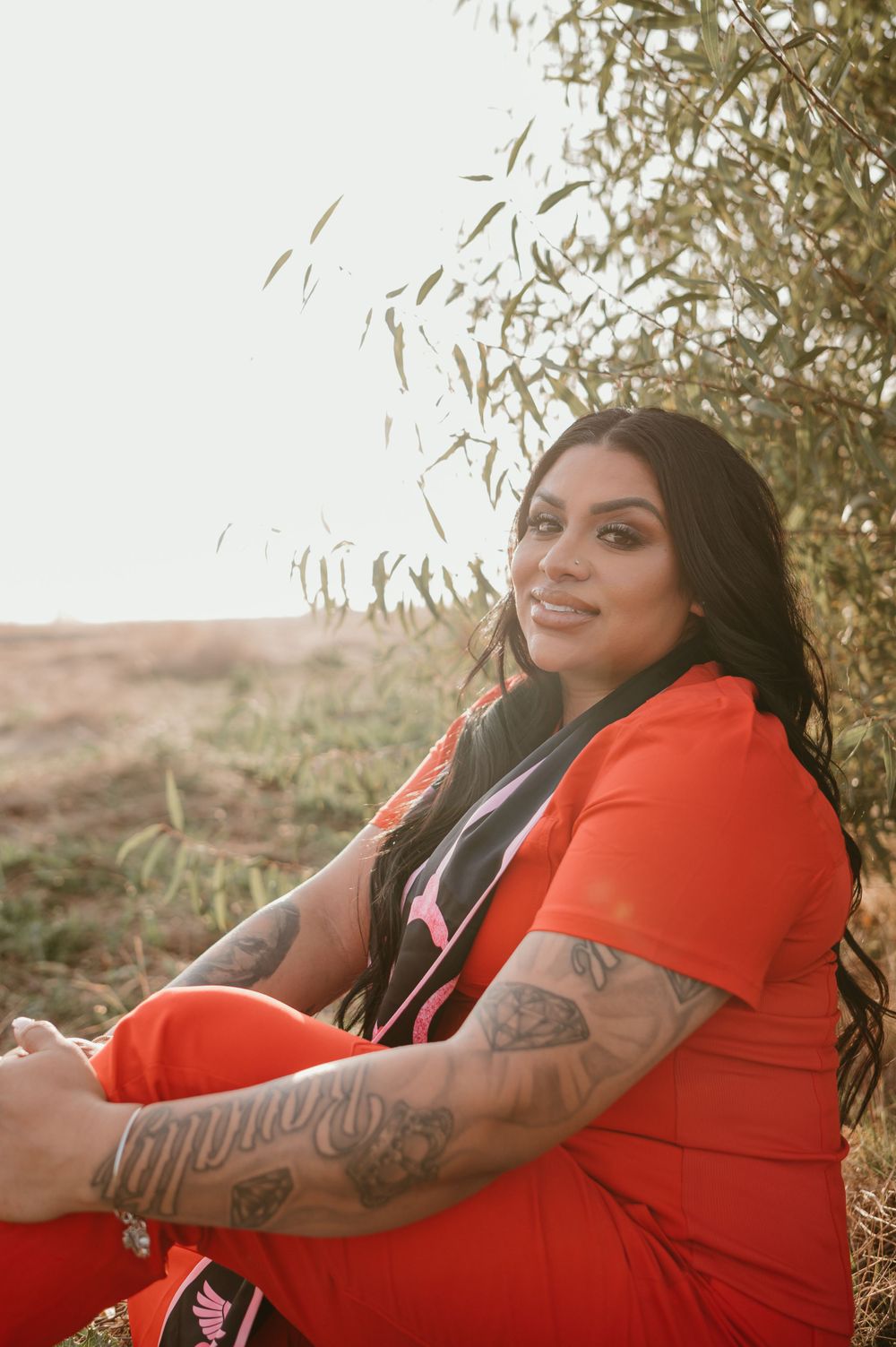 Graduate in red dress shows off arm tattoos while posing in desert setting.