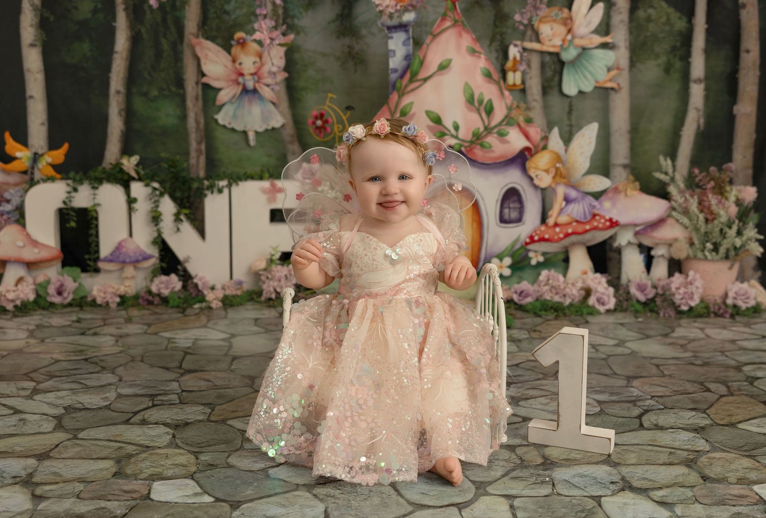 A one-year-old in a peach lace dress sits in a whimsical garden setting with fairy decorations for a birthday photoshoot.