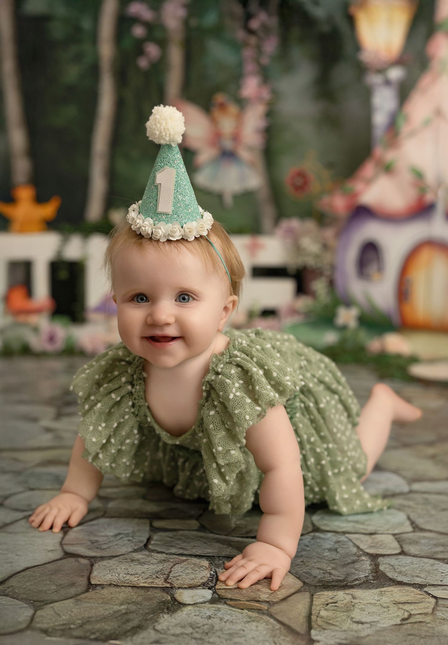 Baby wearing sage green dress and mint party hat crawls on stone patio with fairytale backdrop and pink flowers.