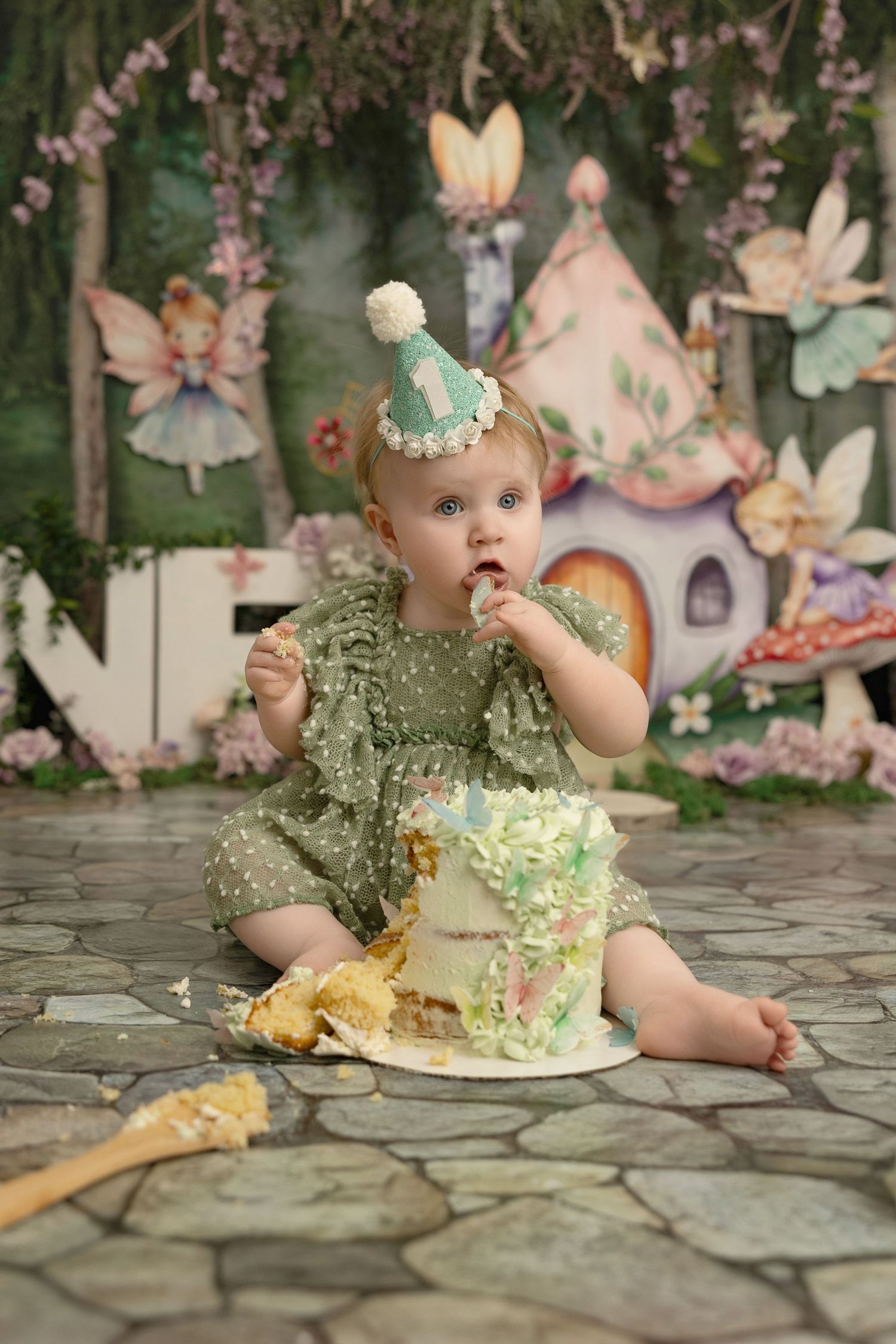 Baby in mint green dress enjoying a cake smash photo session in a whimsical fairy garden setting with pink blossoms.