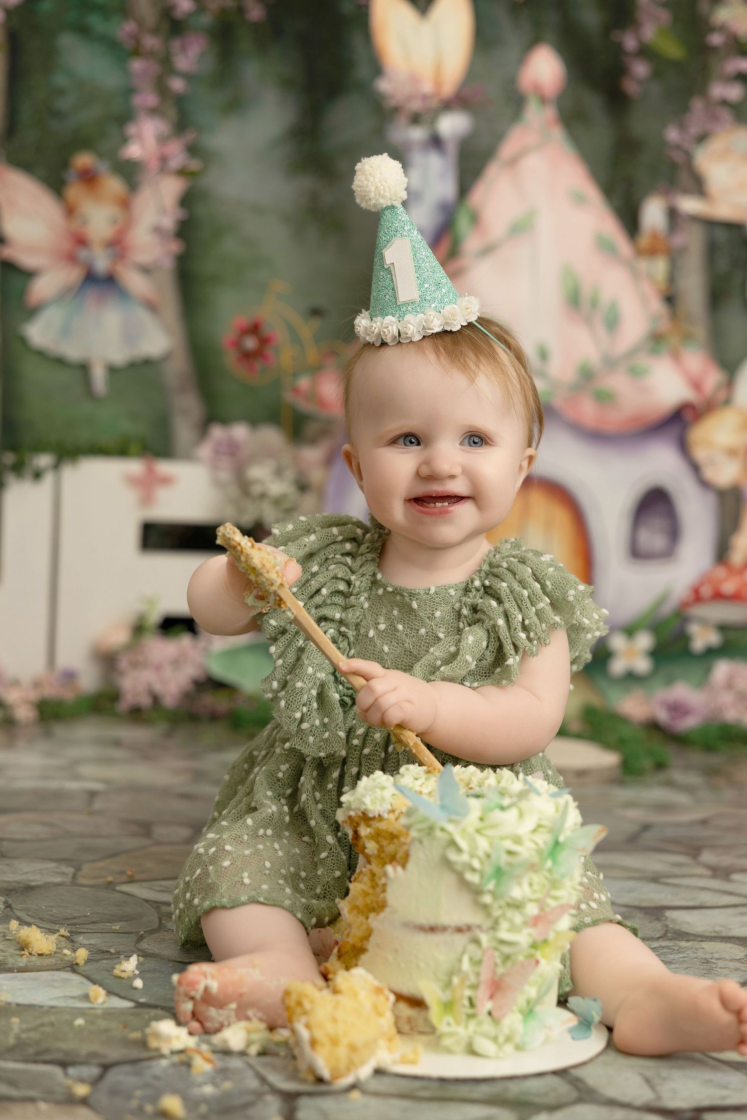 A cake smash photo session featuring a small baby in a mint green dress and party hat enjoying a birthday cake.