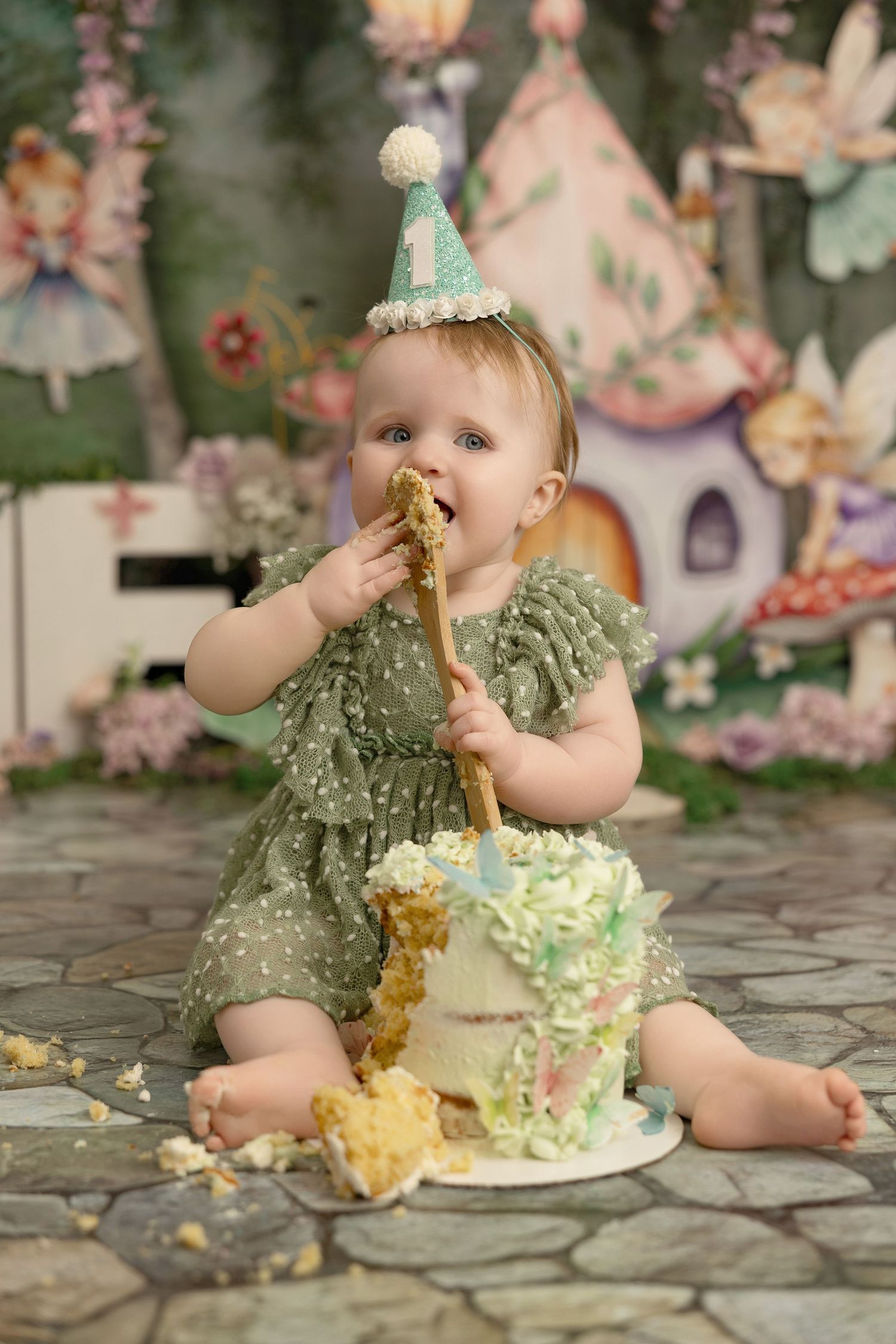 A baby wearing a lace dress sits on a stone patio excitedly eating and smashing their first birthday cake.