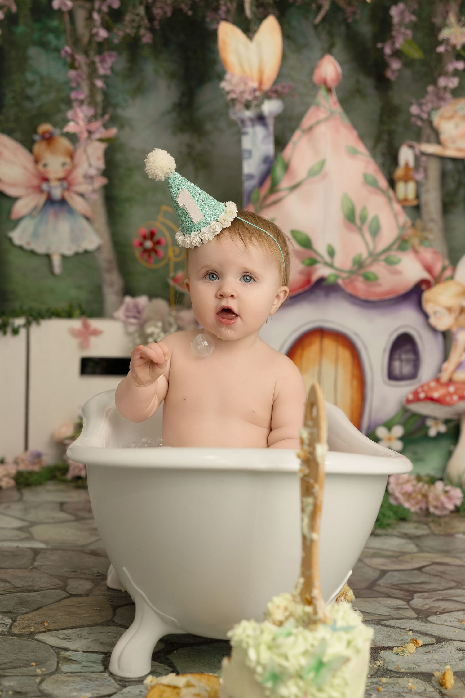 A whimsical bath photo session with a small baby in a party hat sitting in a vintage-style tub with a cake nearby.