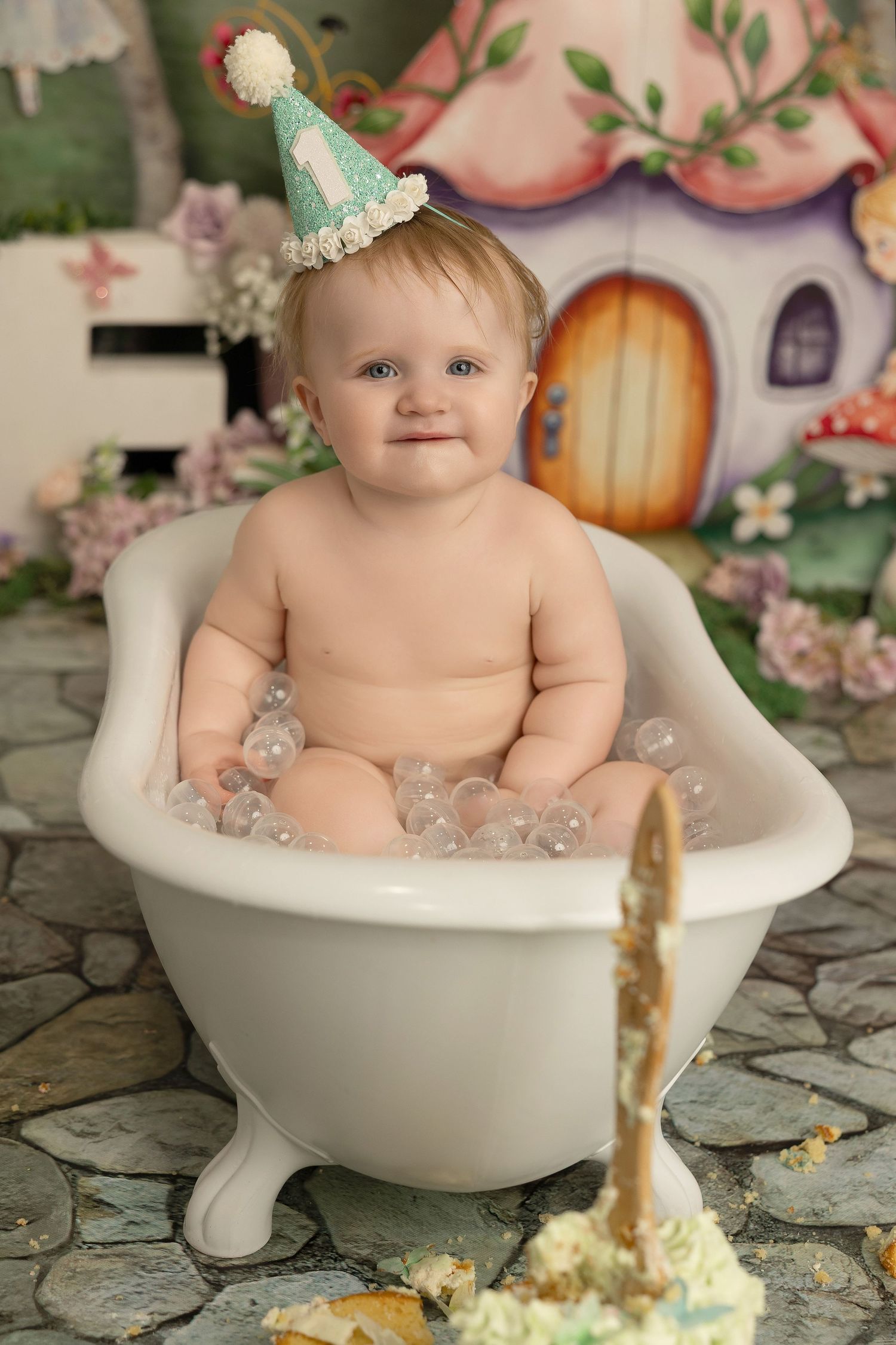 Whimsical bath photography setup with tiny mint party hat, clawfoot tub, and fairy tale cottage backdrop with flowers.