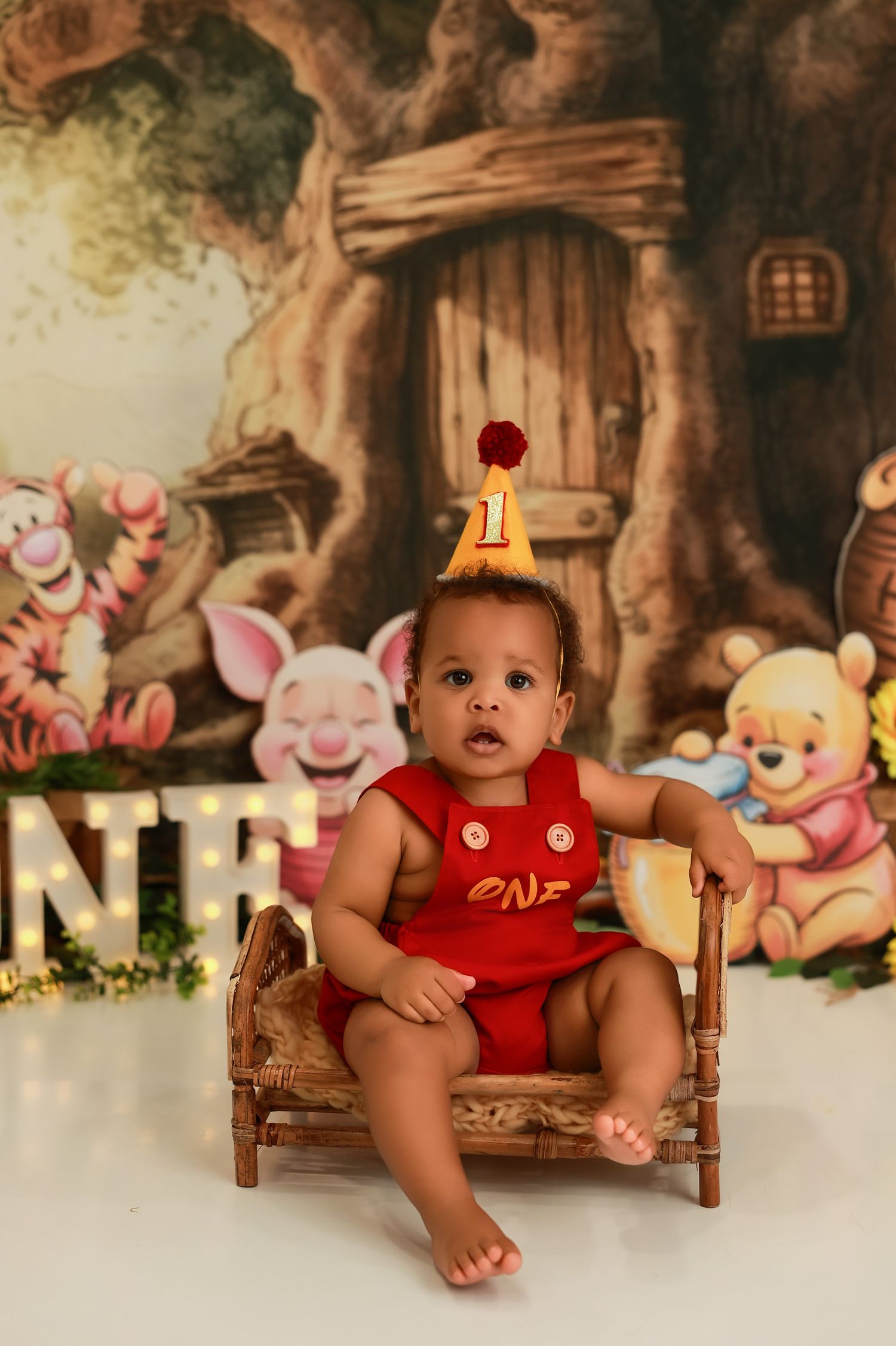 Baby in red outfit sits on wooden bench against whimsical woodland backdrop with stuffed animal decorations.