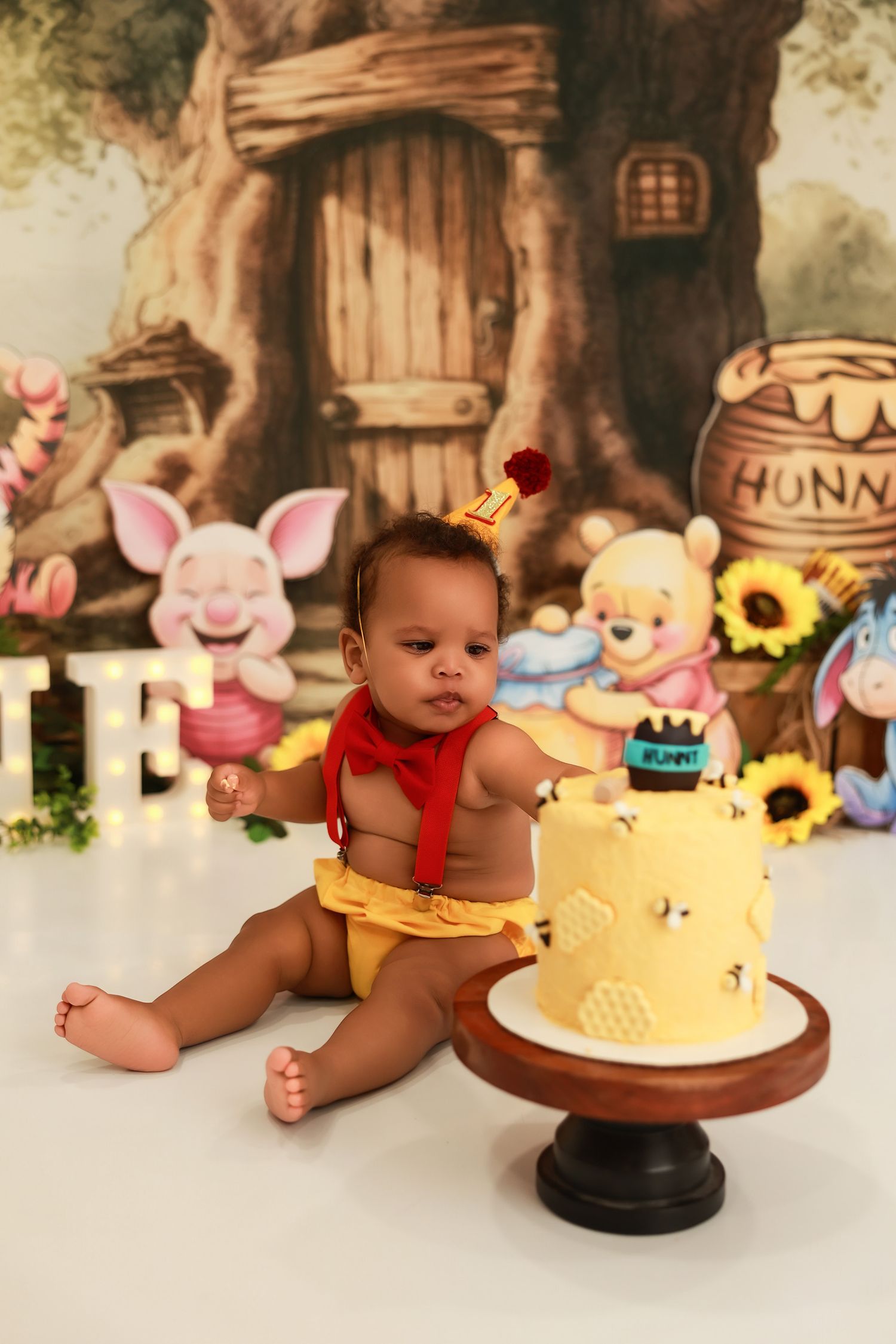 Baby explores yellow birthday cake while seated against fairy tale forest backdrop with plush toys.
