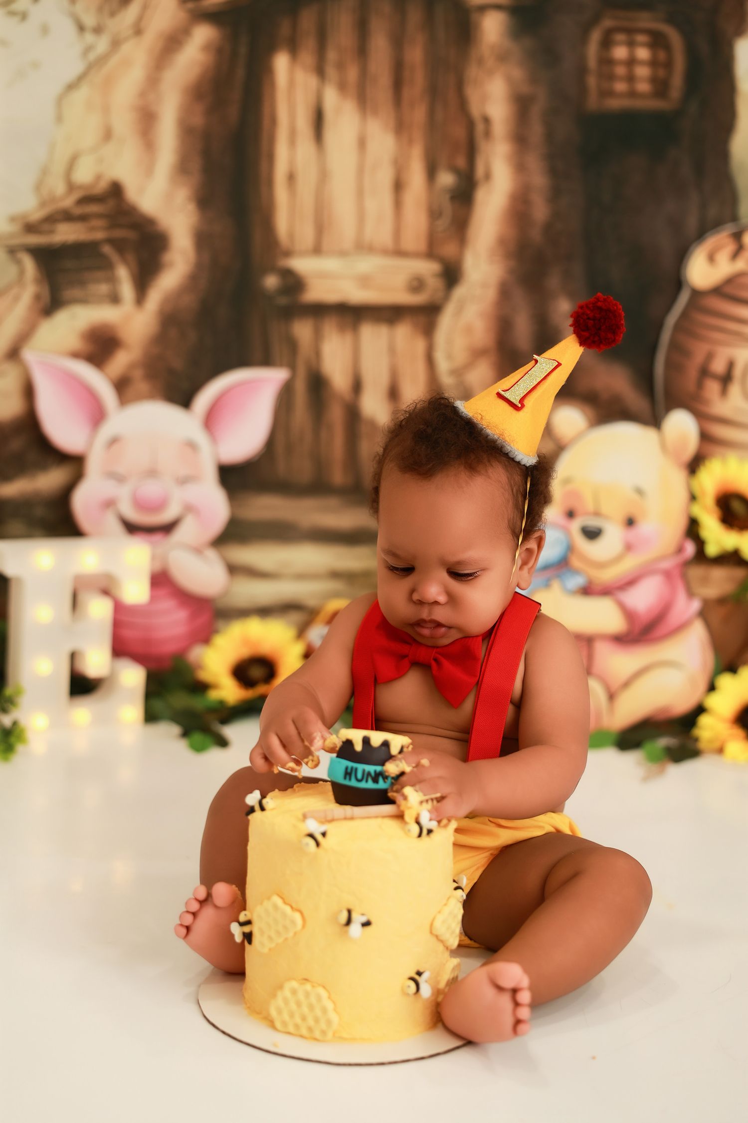 A baby in red outfit explores their first birthday cake with Winnie the Pooh themed decorative backdrop.
