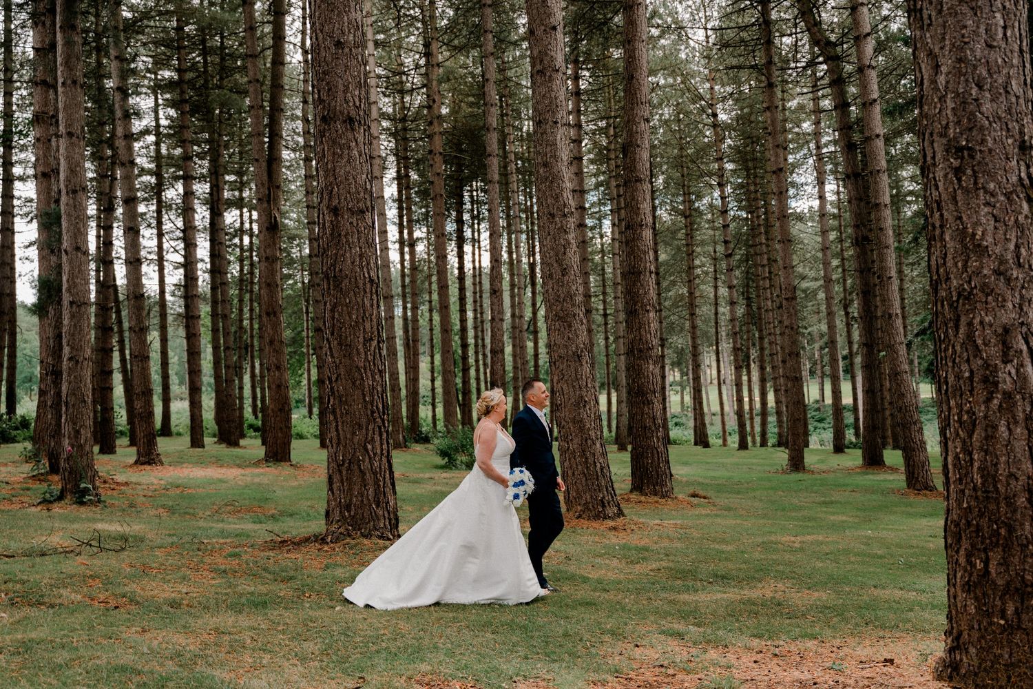 A romantic wedding couple embraces in a serene pine forest with tall trees and green grass.