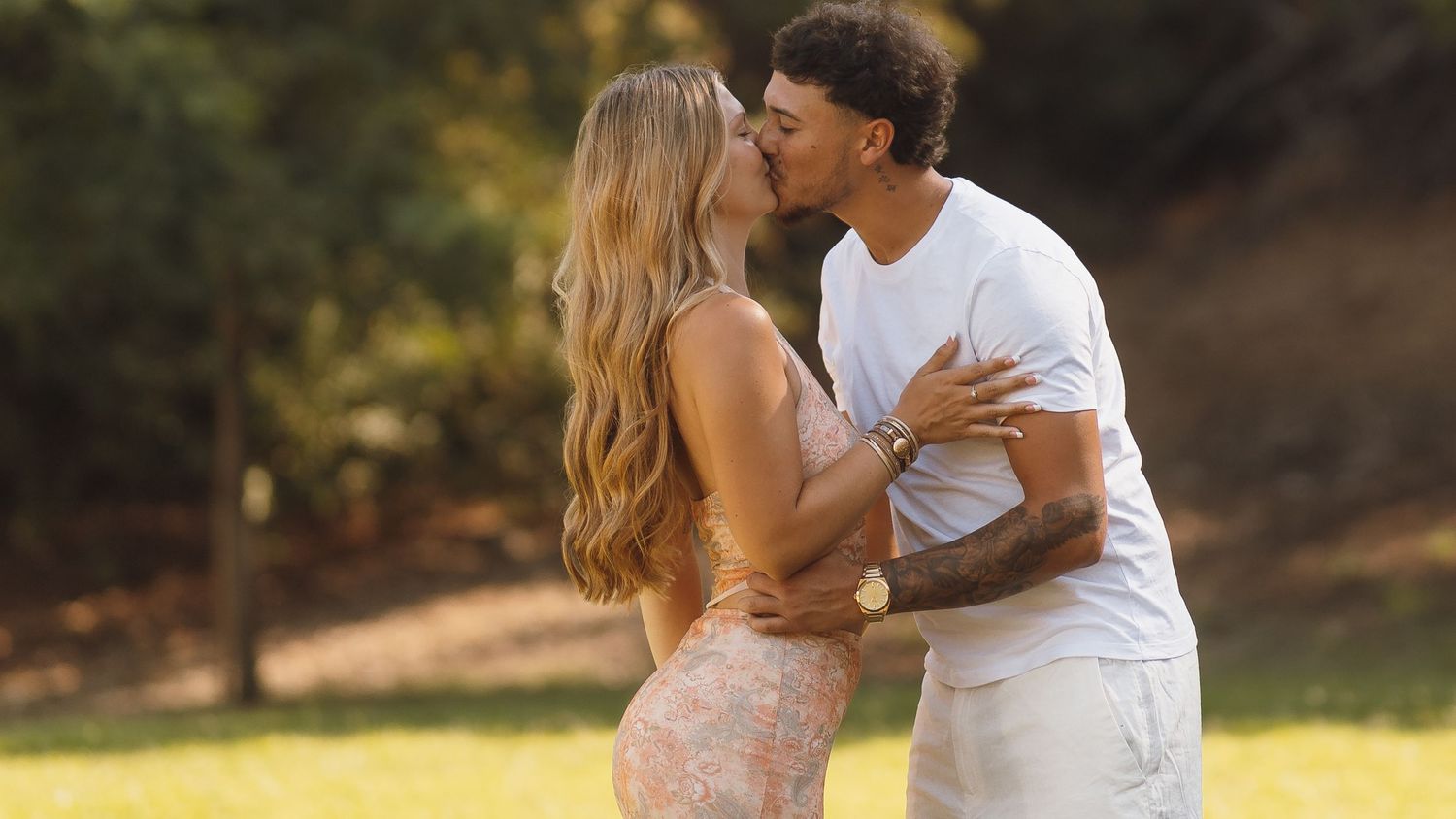 Couple sharing a kiss in a sunlit park, woman in floral dress, man in white outfit.
