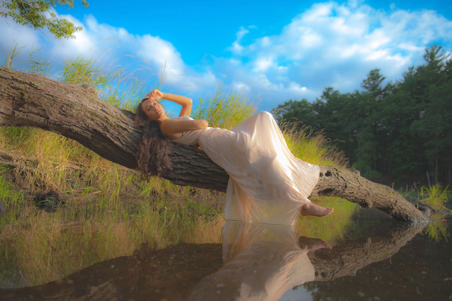 A flowing white dress catches sunlight as someone poses gracefully on a fallen log over water.