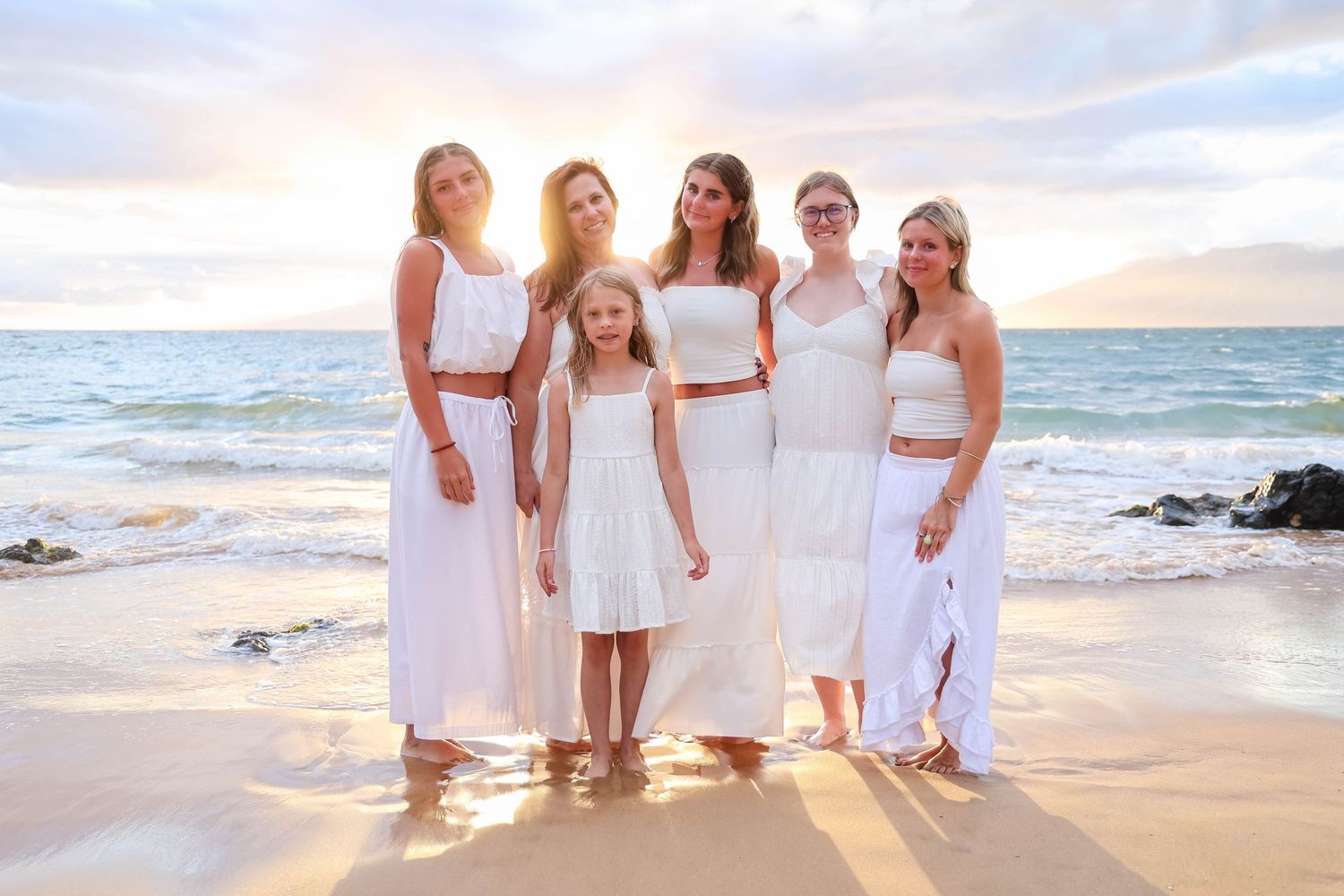 A group in white dresses walks along a scenic beach at sunset while waves crash on the shore.