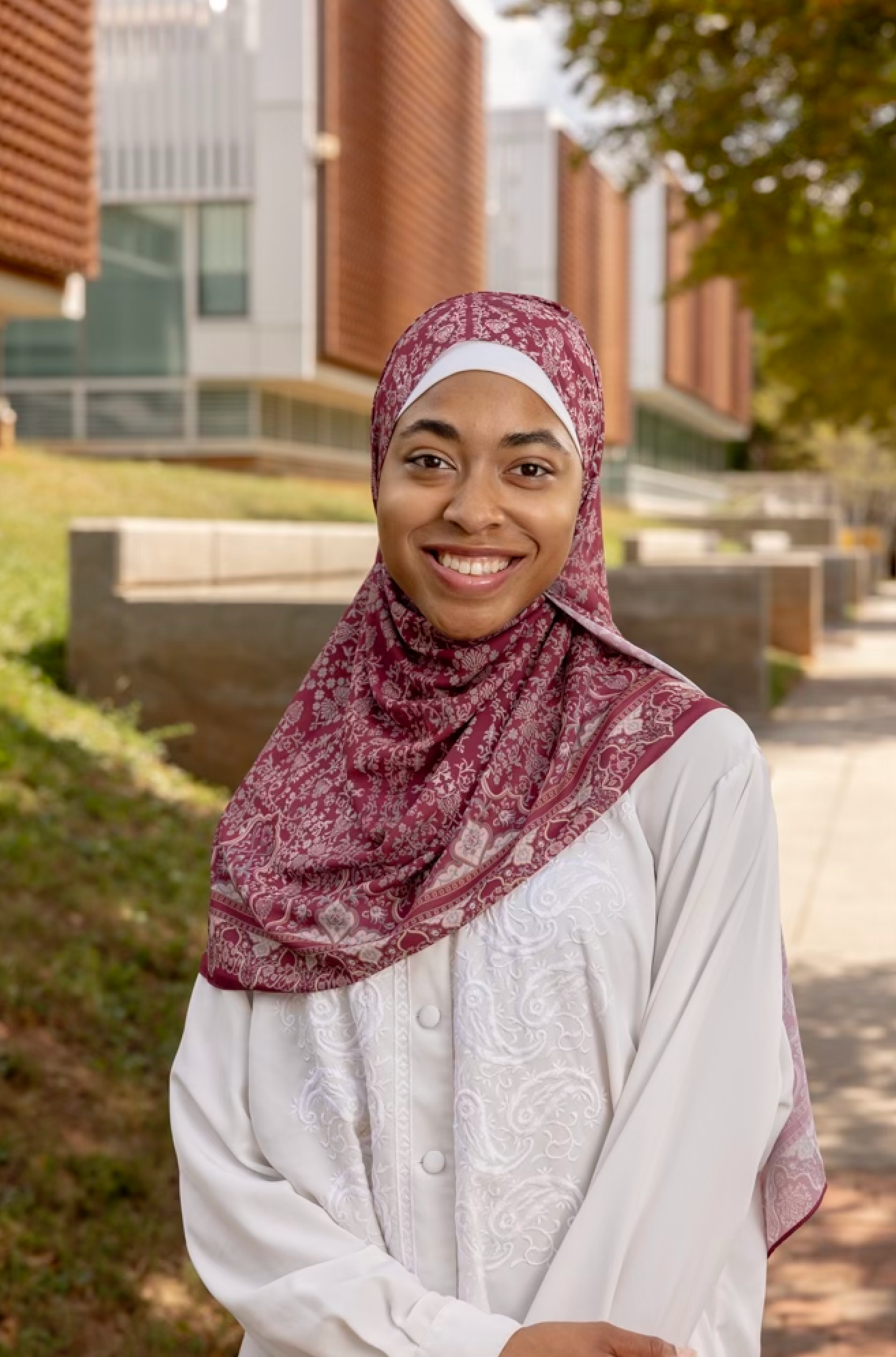 Smiling young woman wearing a maroon hijab and white blouse standing outdoors on a college campus.