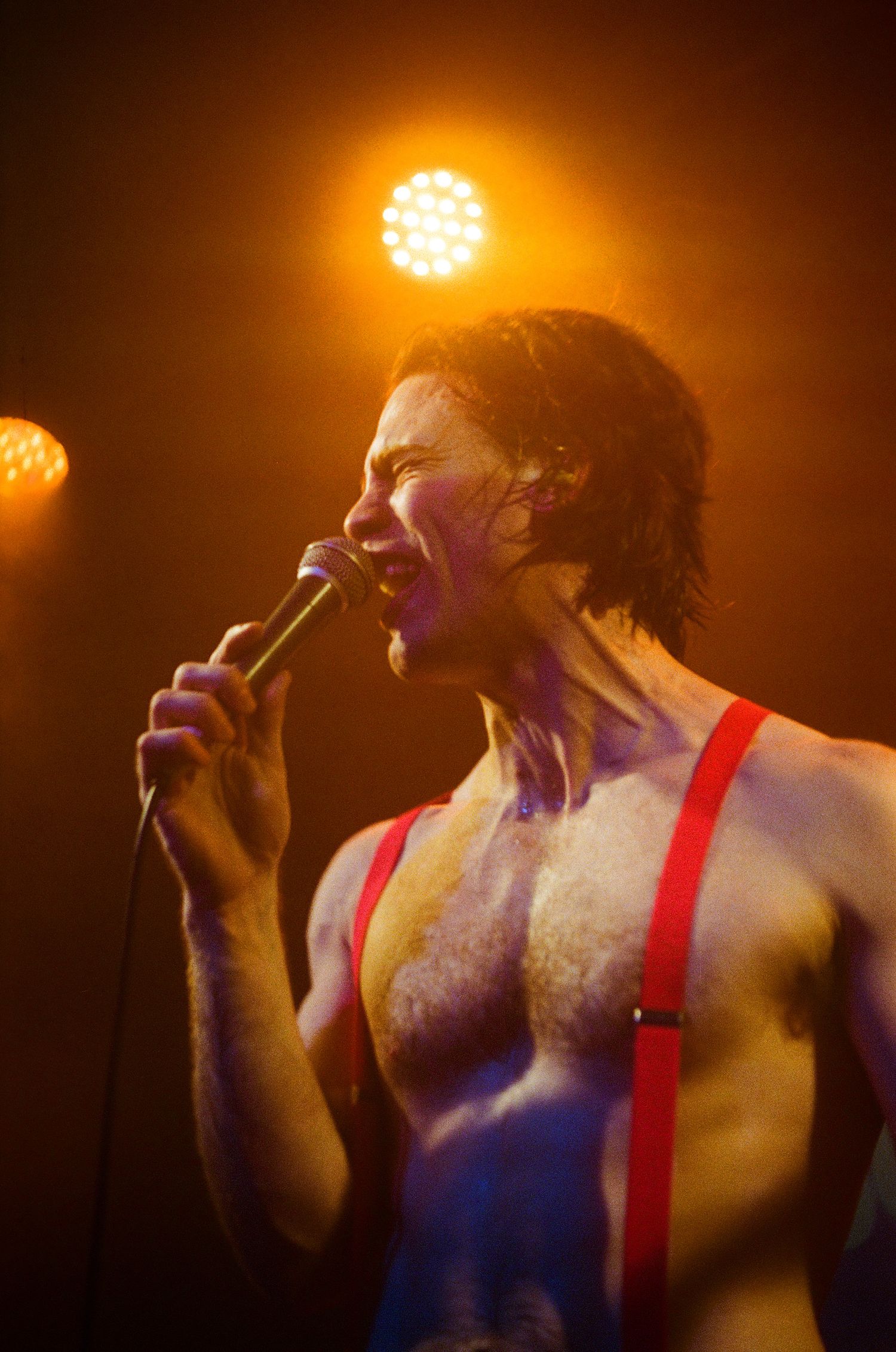 Performer singing passionately under orange stage lights wearing red suspenders.